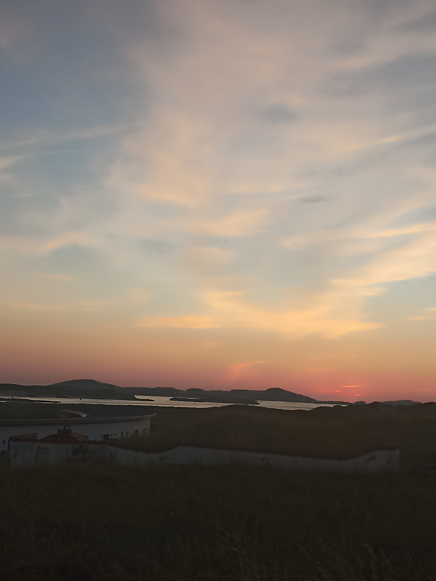 An atmospheric landscape photograph captures a sunset over a body of water and distant hills. The sky is painted with hues of blue, pink, and orange, transitioning from a pale blue at the top to a fiery orange and red near the horizon. Wispy clouds are scattered across the sky, catching the last rays of the sun. In the foreground, a low, circular structure with a partially visible roof is situated on a grassy knoll. The structure appears to be old and possibly abandoned, with some vegetation growing on its roof. Beyond this structure lies the water, reflecting the colors of the sunset, and further in the distance, rolling hills are silhouetted against the vibrant sky. The overall impression is one of tranquility and natural beauty.