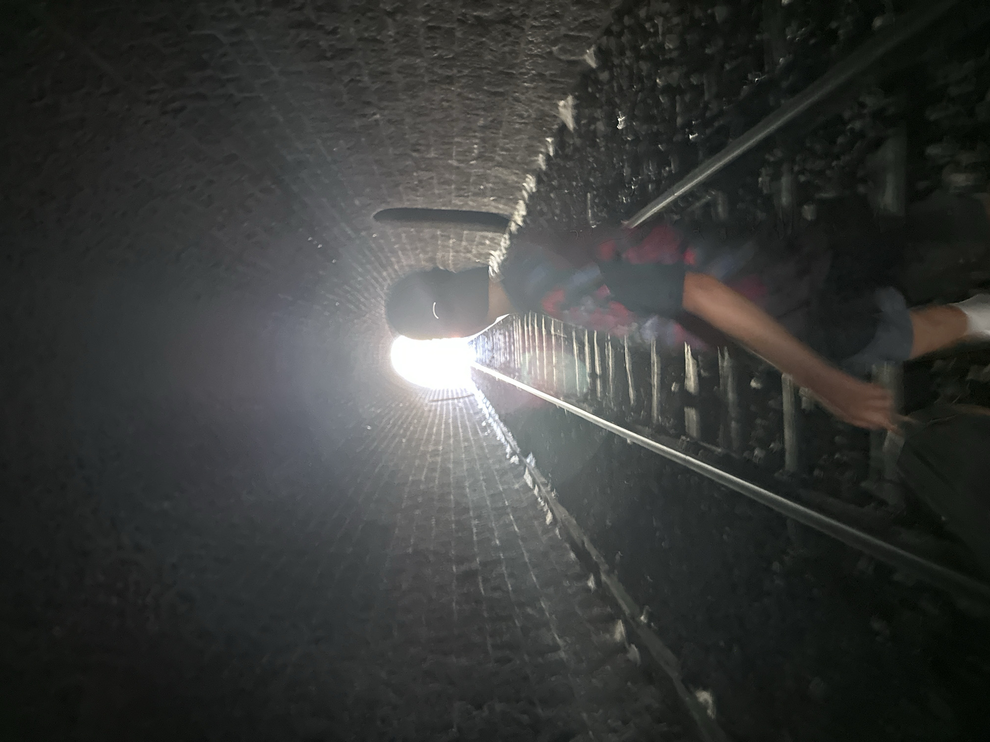 The image shows a person standing on railway tracks inside a stone tunnel. The person is facing away from the camera, towards a bright light at the end of the tunnel. The tunnel walls are made of rough-hewn stones, and the railway tracks are covered in ballast. The scene appears to be located in the Mont Bouquet tunnel in Générargues, France, which is known for its abandoned railway line and has become a popular spot for urban exploration. The person is wearing a t-shirt and shorts, and has a baseball cap on. The lighting is dim, with the bright light at the end of the tunnel creating a high contrast.