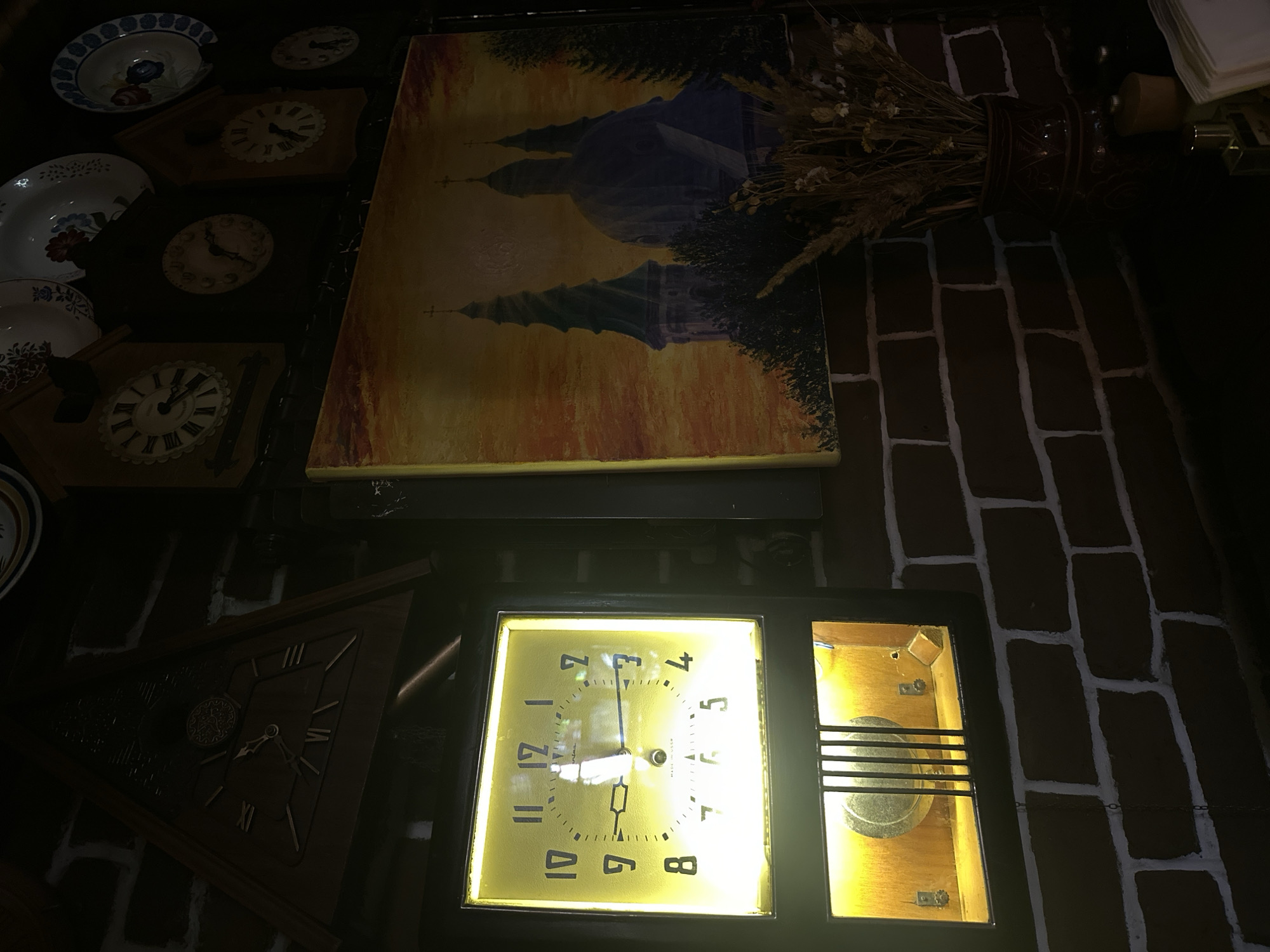 The image depicts a dimly lit interior scene, likely a cafe or a home, adorned with various clocks and artwork. The dominant features include a large, illuminated clock with a yellow backlight, displaying Arabic numerals. Below this clock is a vintage radio with a visible speaker grille. To the right of the illuminated clock, a painting of church spires against a sunset sky is mounted on a brick wall. The brick wall itself is a prominent element, with contrasting dark bricks and white mortar. Several other clocks, of varying designs and sizes, are visible on the wall above the painting, some with Roman numerals. Decorative plates are also displayed on the wall. In the foreground, on a dark surface, a vase filled with dried plants and a few small bottles are present. The overall ambiance is vintage and somewhat rustic. The presence of "MADE IN USSR" on the illuminated clock suggests a connection to the Soviet era, which aligns with the cultural context of Ternopil, Ukraine, a city with a rich history that includes periods under Soviet rule. However, the specific location within Ternopil cannot be determined from the image alone; it could be a historical building, a themed establishment, or a private residence.
