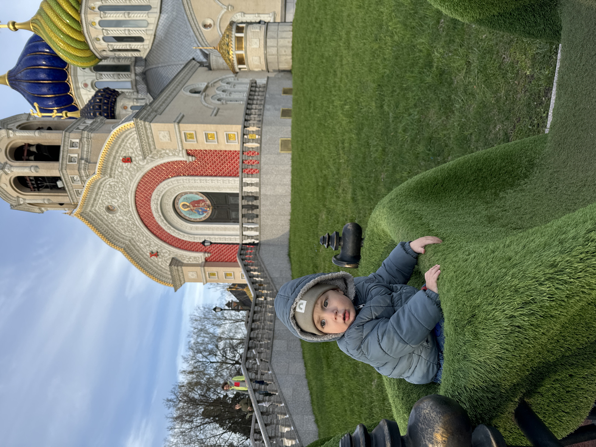 A child is playing on an artificial green surface in front of a building with Russian-style architecture. The building has a large central facade with an arched doorway topped with a red and white patterned design. Above that are golden domes and a blue, onion-shaped dome with golden detailing. There are bare trees in the background, and a blue sky with light clouds. The scene is likely in Moscow, Russia.