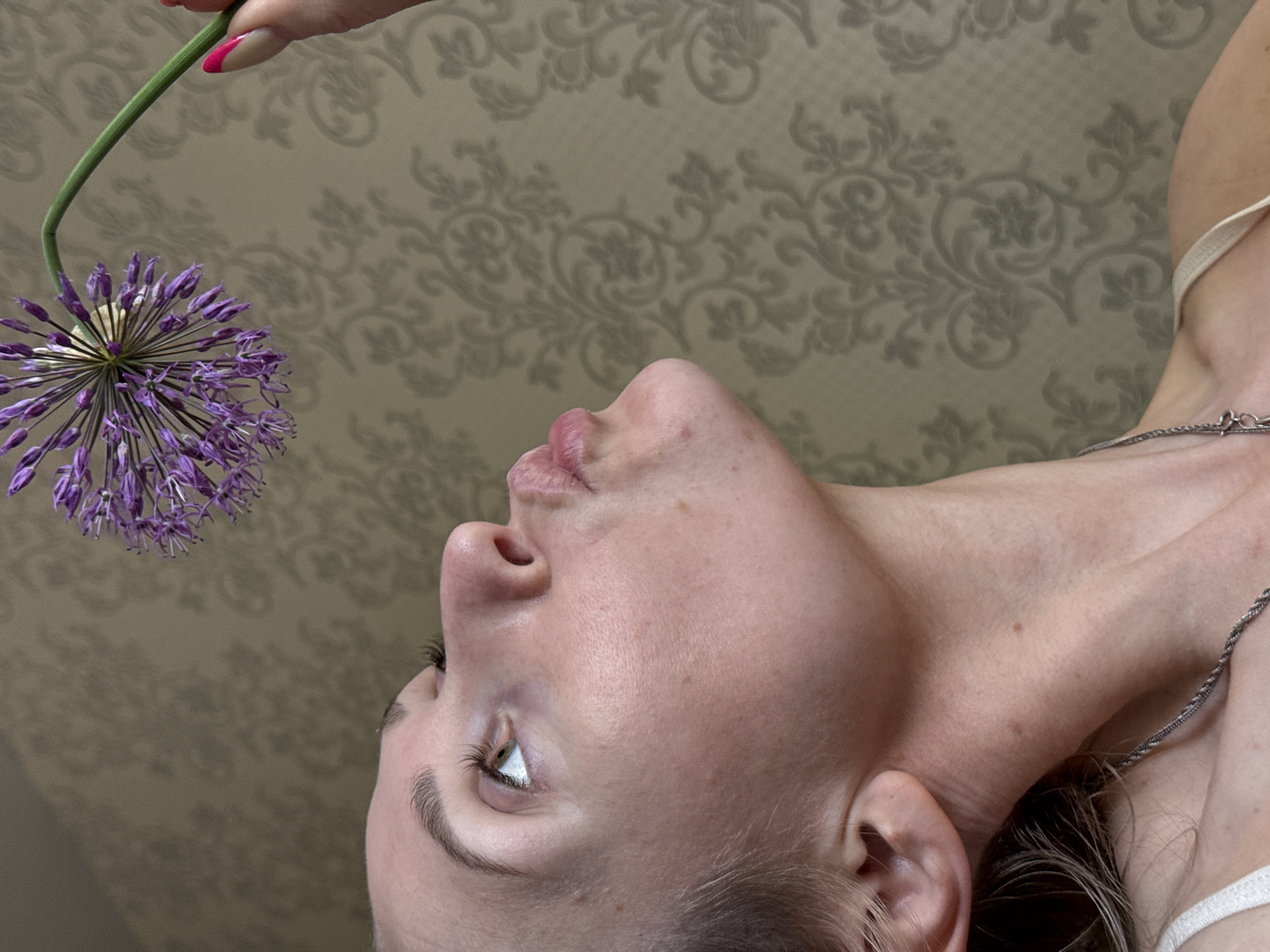 A woman lies on her back, looking up at a purple allium flower held by a hand with a pink-tipped nail. The background is a patterned wallpaper in light beige tones. The woman has fair skin, light brown hair, and a silver necklace. Her expression is one of contemplation. The exact location in the city of Chaikovsky, Russia, is unknown.
