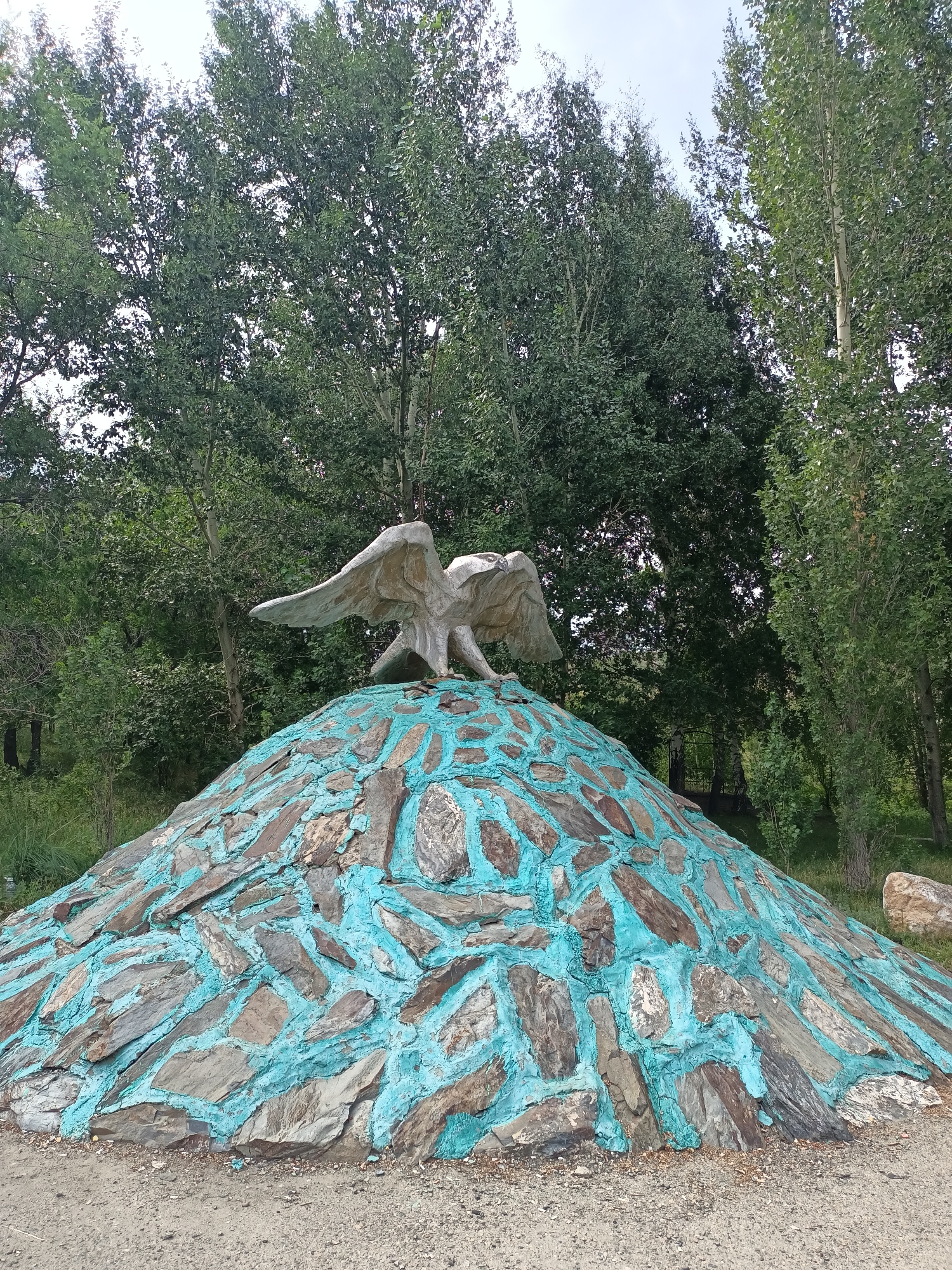 An outdoor, eye-level, medium shot captures a stone sculpture of an eagle with outstretched wings, perched atop a conical mound. The mound is covered with irregular, flat stones, some of which are painted with bright turquoise grout, creating a mosaic-like effect. The eagle sculpture, made of a lighter colored material, is detailed, with visible feathers and talons. Behind the sculpture, a dense backdrop of lush green trees dominates the scene, with a glimpse of a pale, overcast sky peeking through the canopy. The ground in the foreground appears to be a mixture of dirt and gravel. The location is identified as Izgutty Ajtykov, Kazakhstan.