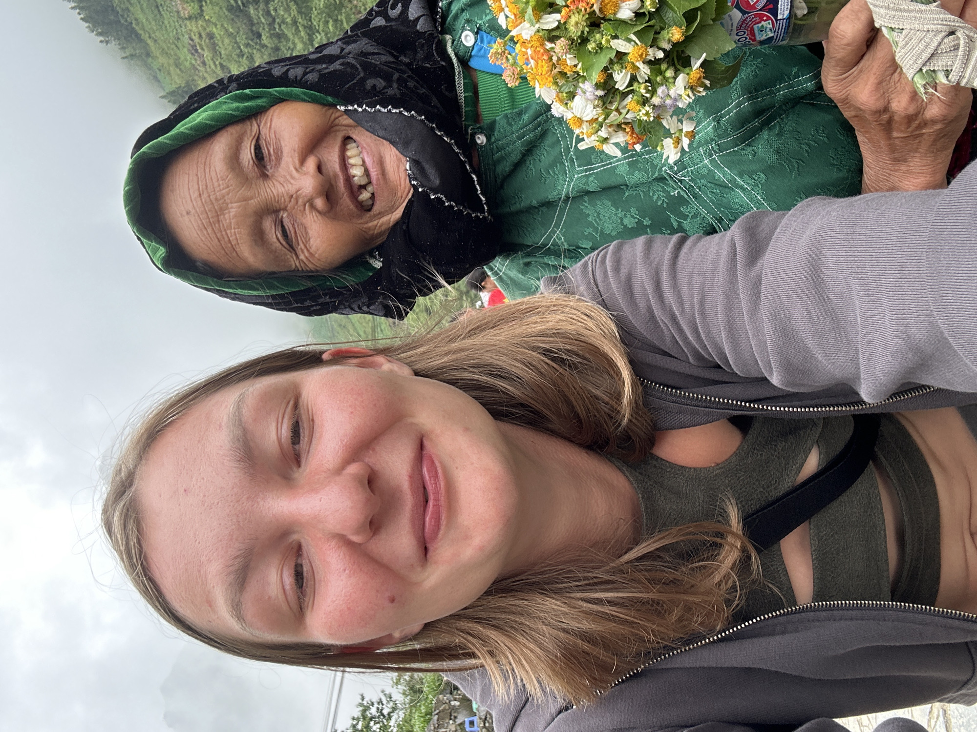 A selfie taken in Yên Minh Commune, Vietnam, shows a young woman with blonde hair wearing a gray zip-up jacket over a dark green top, smiling at the camera. Behind her, an elderly woman in traditional Hmong attire smiles broadly. The elderly woman wears a black head covering with green trim and a dark green jacket with white stitching details and a blue button. She holds a small bouquet of colorful wildflowers. The background reveals a cloudy, mountainous landscape.