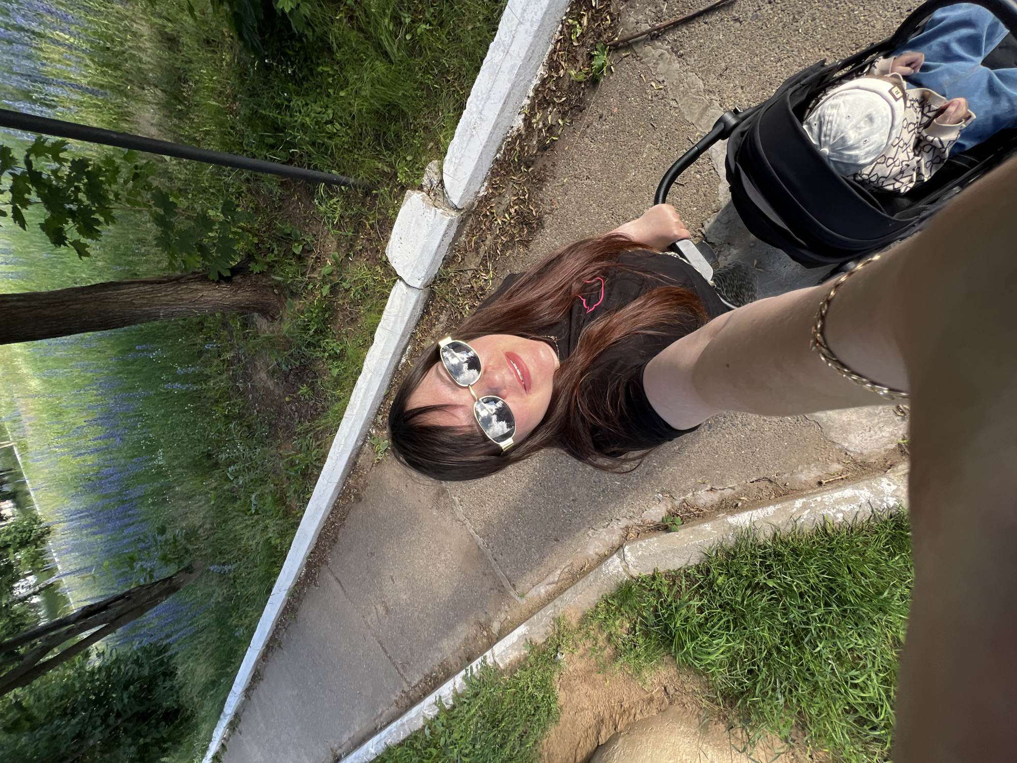 This media file features a woman taking a selfie in an outdoor setting. The woman has long, dark brown hair, is wearing sunglasses, and a black t-shirt with a pink outline of a cat on it. Her left arm is visible, adorned with a bracelet. Next to her, a baby is visible in a stroller, wearing a light-colored cap and a patterned sweater. The scene is set on a paved path bordered by a concrete curb. To the left of the path, there's a grassy area with trees and a field of blue flowers, suggesting a park or natural landscape. The lighting indicates it's daytime, likely sunny given the reflections in the woman's sunglasses. The overall mood appears casual and relaxed, possibly a walk in the park with the baby.