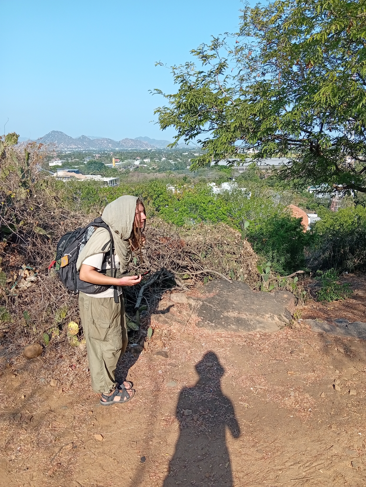 The image presents a person standing on an arid hillside, overlooking a town and mountains under a clear, bright blue sky. The individual, appearing to be an adult with dreadlocks, wears a light-colored head covering, a t-shirt, olive green cargo pants, and sandals. They are equipped with a dark backpack, suggesting a hiking or travel activity, and hold a small, dark object in one hand.
The foreground consists of dry, reddish-brown earth with sparse vegetation, including tangled dried branches and thorny bushes, possibly cacti. A large rock is visible on the ground to the right. A prominent shadow of the photographer is cast on the ground in the lower foreground, indicating strong, direct sunlight, likely mid-morning or mid-afternoon.
Beyond the immediate foreground, a sprawling town or city unfolds, characterized by numerous buildings nestled among abundant green trees. In the distance, a range of mountains rises against the horizon. A large tree with green leaves and some visible pods or fruit partially frames the right side of the scene. The weather is sunny and clear, indicative of a warm climate. The overall setting aligns with an elevated viewpoint in an area like Phan Rang – Tháp Chàm, Vietnam. The person appears to be observing the expansive landscape.