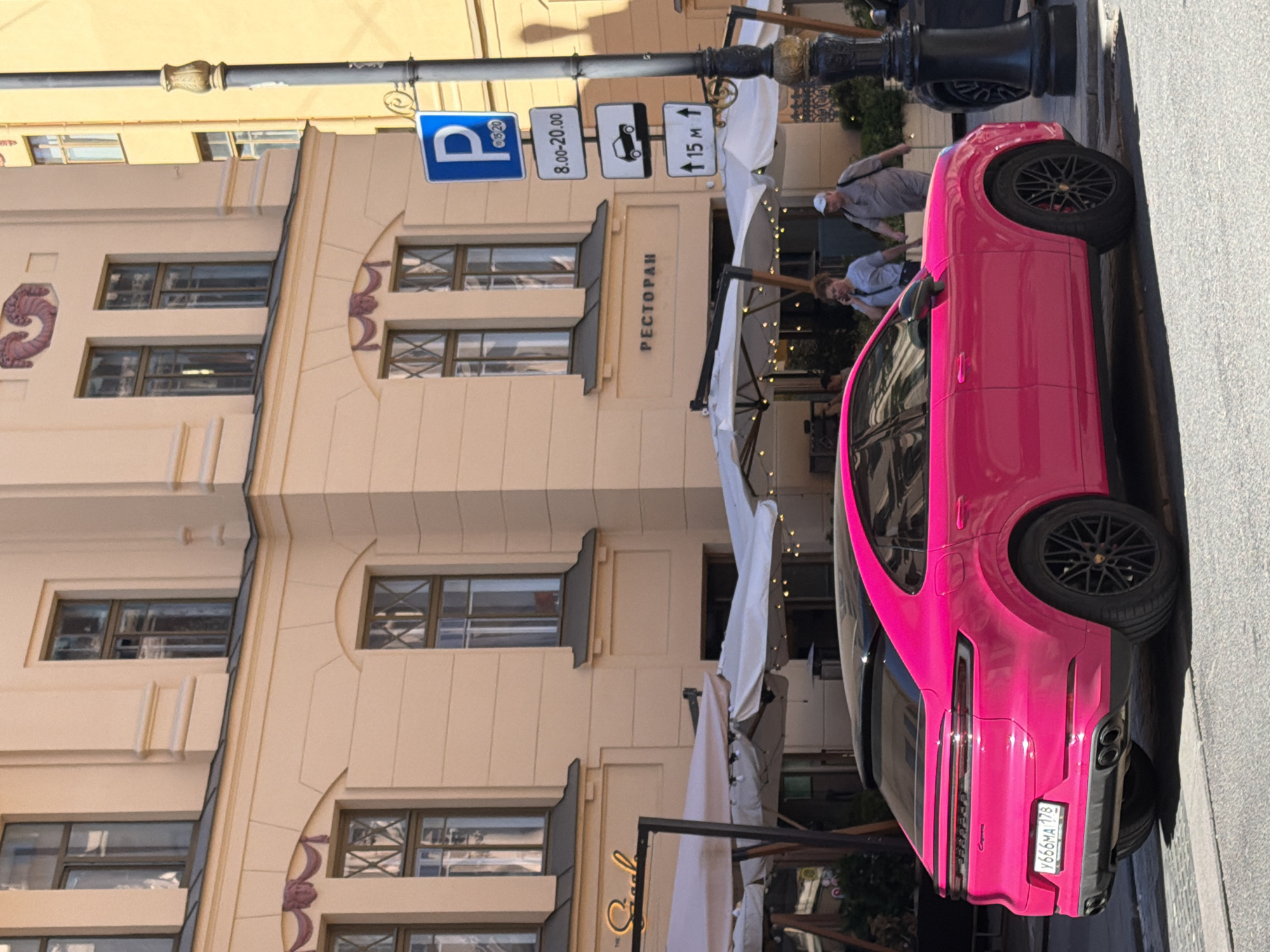 The image depicts a vibrant street scene on a sunny day in Saint Petersburg, Russia. A striking bright pink (magenta) Porsche Cayenne Coupe with black wheels and a black roof is prominently parked on the right side of an asphalt street, facing right. Its Russian license plate, "Y666MA 178", is clearly visible on the rear.
In the background, a multi-story building with a light peach-colored facade and classical architectural details, including ornate window frames, cornices, and decorative relief elements, dominates the left and center. The ground floor of this building features an outdoor cafe or restaurant, indicated by several white umbrellas, string lights, and two signs: one reading "РЕСТОРАН" (RESTORAN - Restaurant) and another partially visible, possibly "SaaL".
Adjacent to the parked car, on the right side of the street, stands a dark grey lamppost with a decorative, gold-toned base. Attached to the lamppost are a series of white and blue traffic signs: a blue square 'P' (parking) sign with "10-15-20" text, a white rectangular sign indicating "8.00-20.00" for parking hours, a white sign with a car icon and a downward arrow, and another white sign with an upward arrow pointing to "15 M".
Two individuals are visible walking on the sidewalk behind the pink car, moving from left to right. A woman with brown hair, wearing a light grey t-shirt and dark pants, is conversing on a dark smartphone held to her ear. Behind her, a man wearing a light t-shirt, light trousers, and a cap walks, carrying a bag. A partial view of a dark-colored vehicle is discernible behind the lamppost on the far right, next to a yellow building. Strong shadows cast across the street indicate direct sunlight.