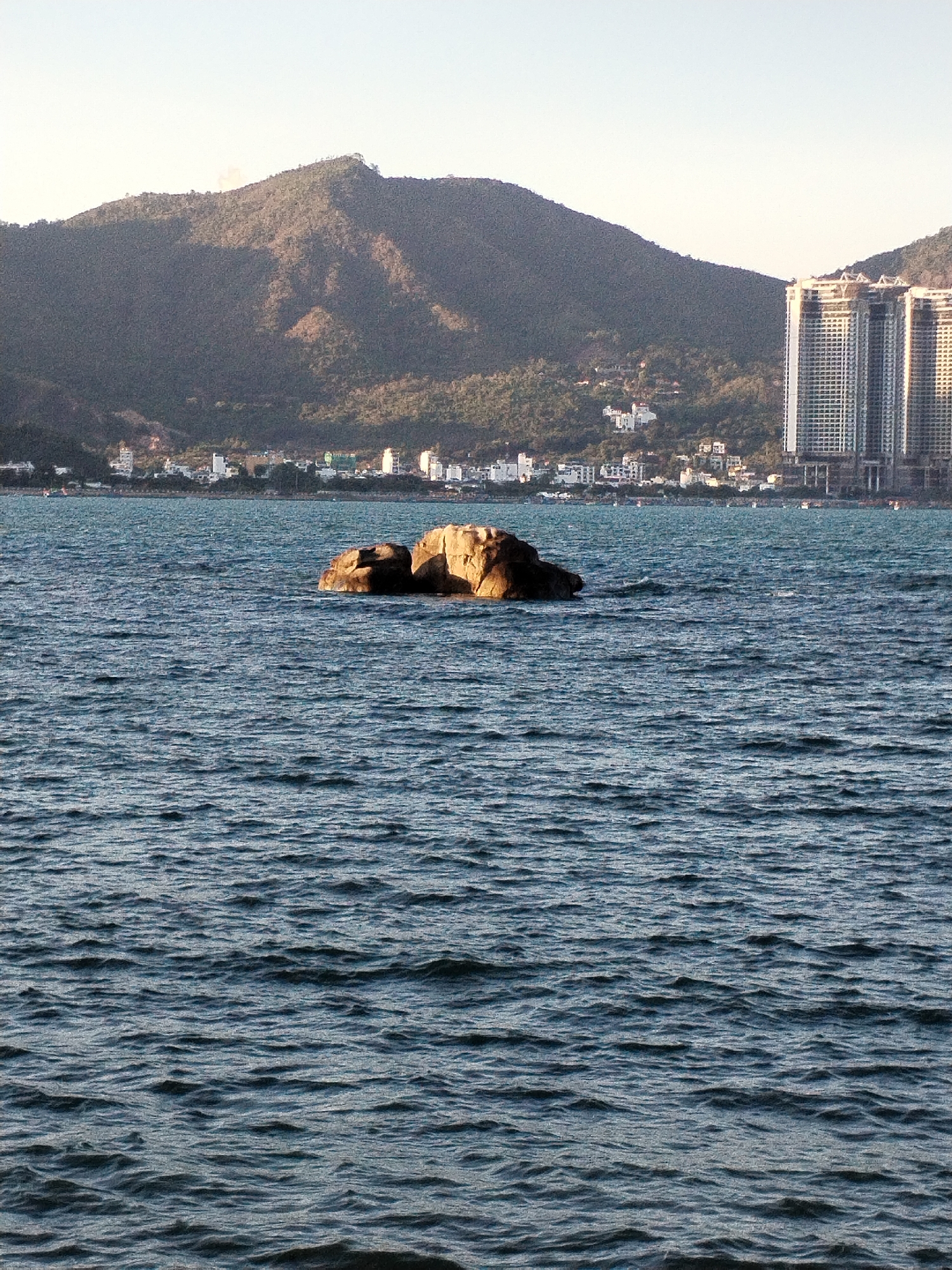 The image shows a rock formation in the water in Nha Trang Ward, Vietnam, with a mountain and buildings in the background. The water is dark blue and choppy. The rock is brown and appears to be partially submerged. The mountain is covered in trees and stretches across the horizon. Some buildings are located on the shoreline and others are taller and located on the mountain slopes behind the town. The sky is clear.