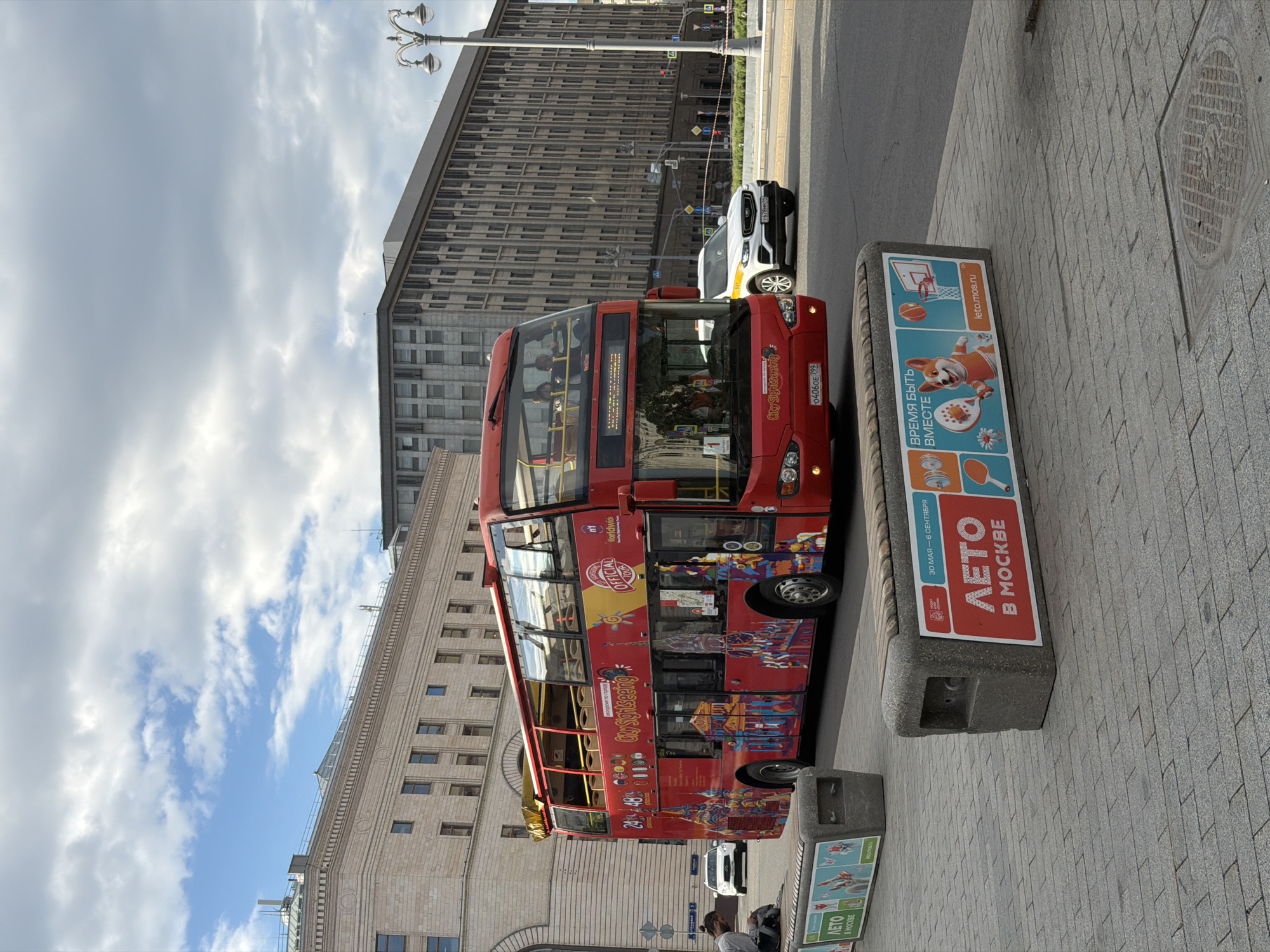 The image depicts a red, double-decker "City Sightseeing" bus in Moscow, Russia. A white SUV is parked behind the bus. Buildings with multiple stories are in the background. A bench with "Summer in Moscow" ("ΛΕΤΟ В МОСКВЕ") signage is visible next to the bus on a paved sidewalk. A license plate on the bus reads "04060E 799." There are people on the bus and on the sidewalk. The sky is partly cloudy.