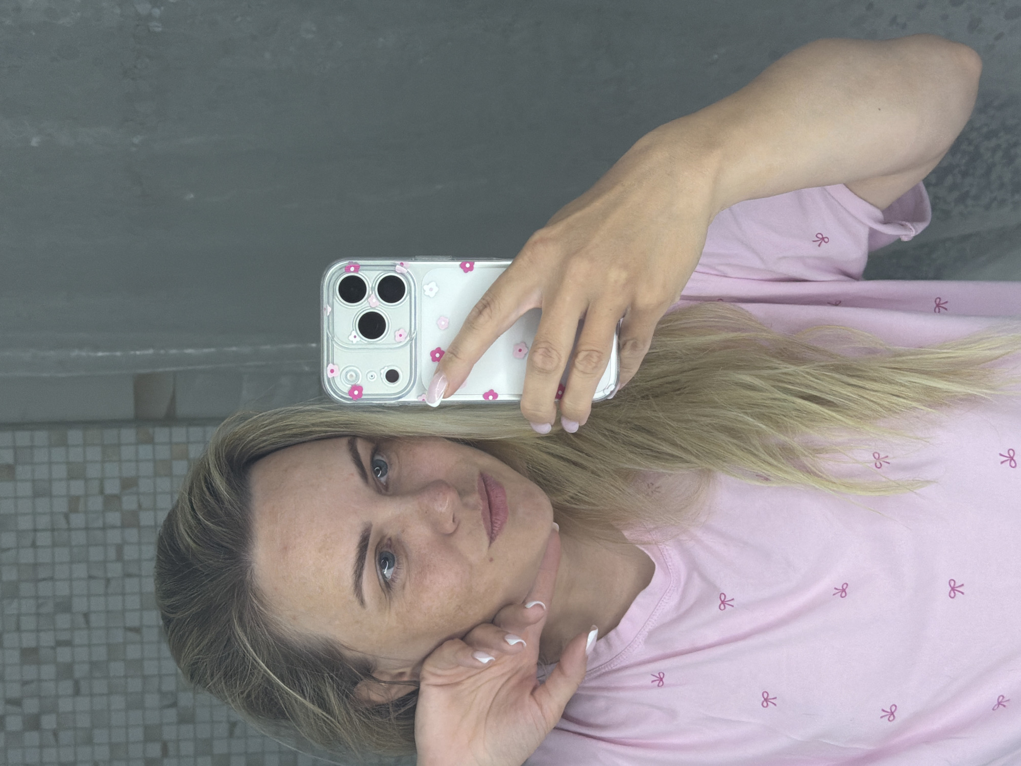 An indoor portrait shows a woman taking a selfie reflected in a mirror or similar surface. She has blonde hair, blue eyes, and light skin with visible freckles, looking directly at the camera with a neutral expression. She is wearing a light pink t-shirt adorned with small, repeating red bow patterns. Her fingernails are painted with a white tip design.
The woman's right hand holds a modern smartphone, positioned near the top of the frame. The phone is encased in a clear cover decorated with small pink and white floral or bow motifs, particularly prominent around its triple camera lenses. Her left hand is gently resting on her chin.
The background behind her head and upper body consists of a dark gray, plain wall or surface. To her left, a light gray tiled wall with small square tiles is visible. The lighting suggests an indoor setting, and there are no discernible location cues within the image itself beyond the interior architecture. The activity depicted is a self-portrait, suggesting an everyday moment. No visible text is present on any objects. The overall scene conveys a casual, personal photograph.