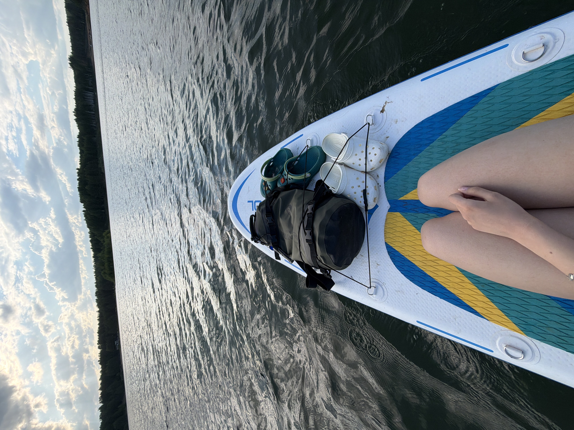 The image depicts a person sitting on a paddleboard in a body of water. The board is white with blue, green, and yellow accents, and has a dark bag, a pair of green Crocs, and a pair of white Crocs secured to the front. The person has their legs bent and their hands resting on their legs. The water is dark and rippled, and the shoreline with trees is visible in the distance. The sky is partially cloudy. It's impossible to verify the location as Selychka, Russia from the image.