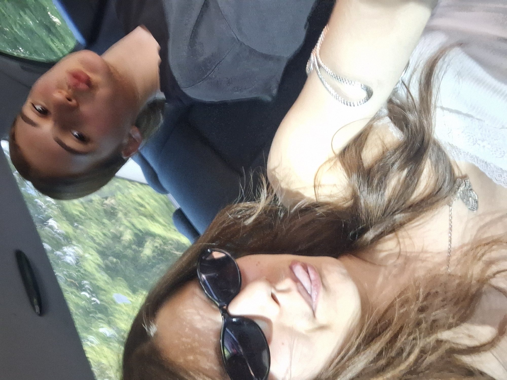 The media file captures a selfie of two young women, likely friends, possibly in a car. The perspective suggests they are lying down, with one woman's face prominently in the foreground, looking towards the camera with an open mouth and partially visible tongue. She is wearing black sunglasses pushed down her nose and a light-colored top with lace detailing. Her long, brown hair is spread around her.
Behind her, another woman's face is visible, turned to the side. She appears to be looking slightly away from the camera, with a subtle expression. She is wearing a dark top. A silver bracelet adorns her wrist. The background shows a blurry green and white landscape, likely foliage seen through a window, suggesting they might be traveling. The lighting is bright, indicating it's daytime, and the overall mood seems casual and relaxed, possibly during a road trip or outing. There is no visible text in the image. The location context provided is Koprivshtitsa, Bulgaria, which might imply the landscape is typical of that region.