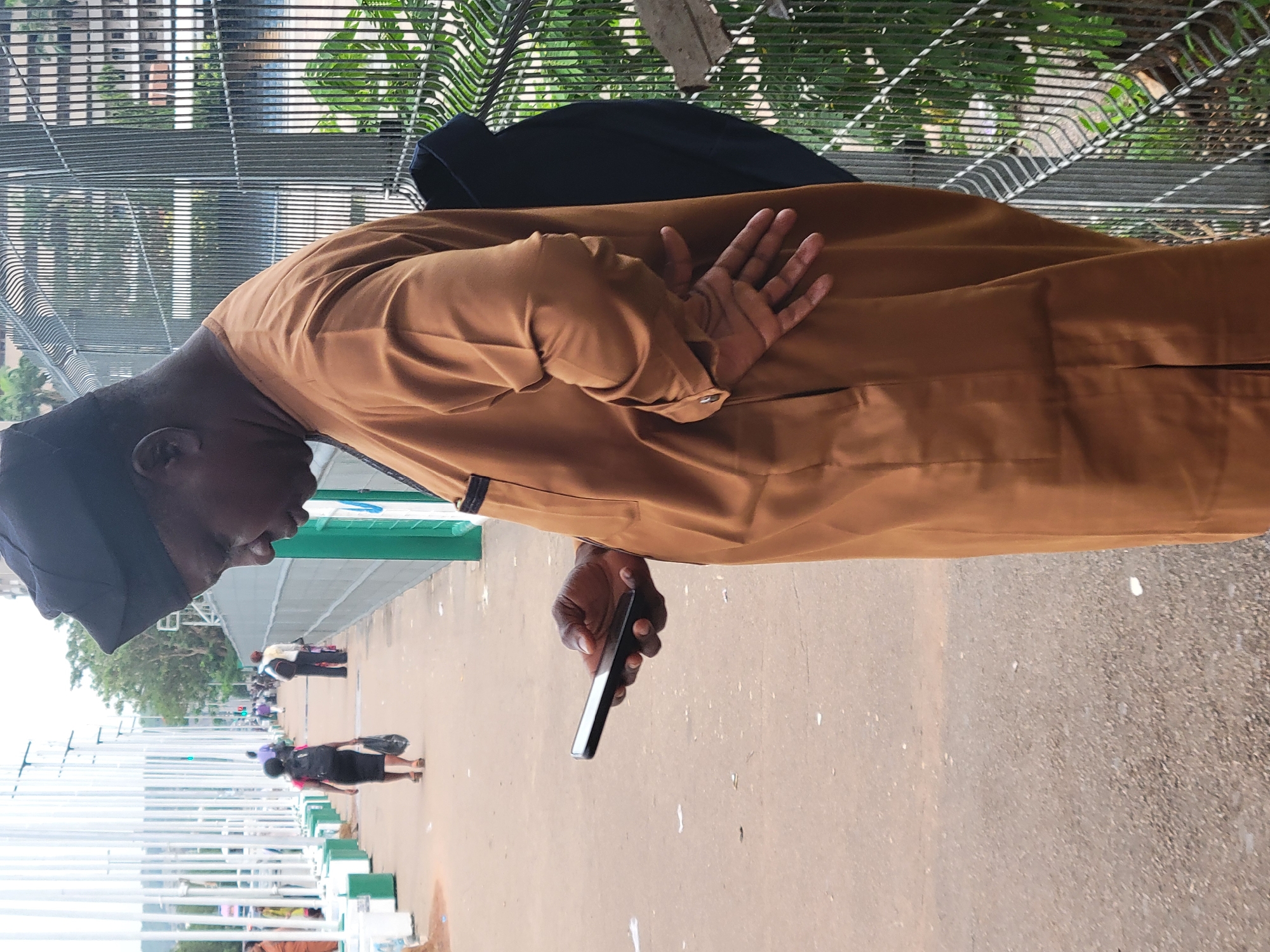 An outdoor, daytime shot, likely taken in Abuja, Nigeria, captures a bustling pedestrian area. The primary subject is a dark-skinned man in the foreground, facing right, dressed in a traditional brown agbada or kaftan and a dark cap. He is holding a black smartphone in his right hand, looking down at the screen, suggesting he is engrossed in its content. His left hand is raised across his chest, with fingers slightly spread.
Behind the man, a metal fence or grille runs horizontally across the mid-ground, with green foliage and distant buildings visible through its pattern, indicating an urban landscape. The ground is a light-colored concrete or paved surface. In the background, several other people are visible. One woman, dressed in a dark top, skirt, and heels, carries a bag and walks away from the camera. Another person in lighter attire also appears to be moving.
The scene features various white and green structures, possibly pillars, barriers, or part of a public facility like a bus station or pedestrian bridge, extending into the distance on the left. The overall impression is that of a typical, busy day in a public space, with people going about their routines. There is no discernible text visible in the image.