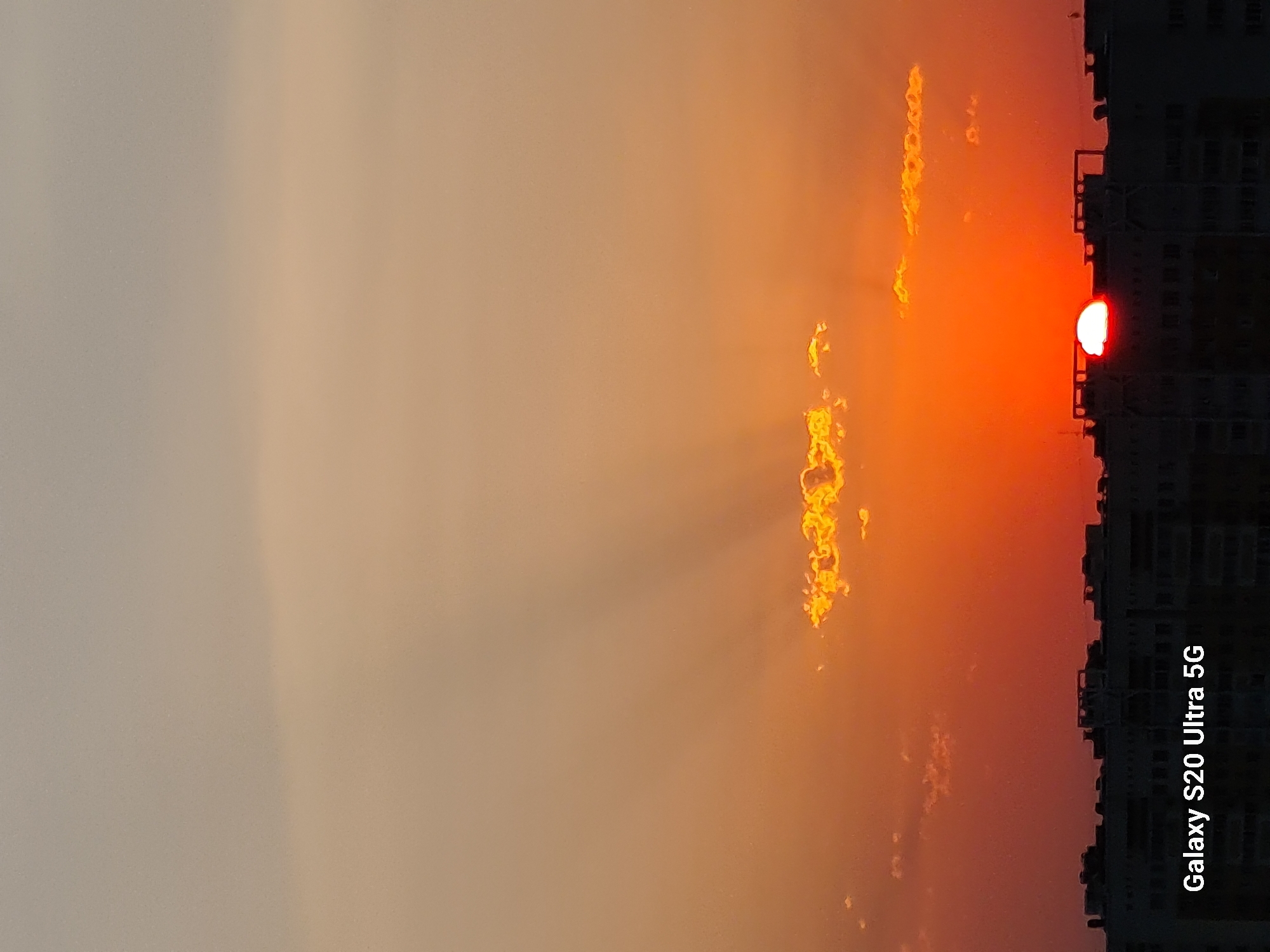 The image depicts a sunset over the city of Almaty, Kazakhstan. The sun, partially obscured by buildings, is a vibrant orange orb low on the horizon. Rays of light emanate from the sun, piercing through a line of clouds. The sky transitions from a pale grey at the top to a deep orange and red near the horizon, indicating the late stage of sunset. In the foreground, the silhouettes of multi-story buildings form a dark line against the colorful sky. A watermark "Galaxy S20 Ultra 5G" is visible in the bottom left corner. The scene is devoid of any people or discernible activity.