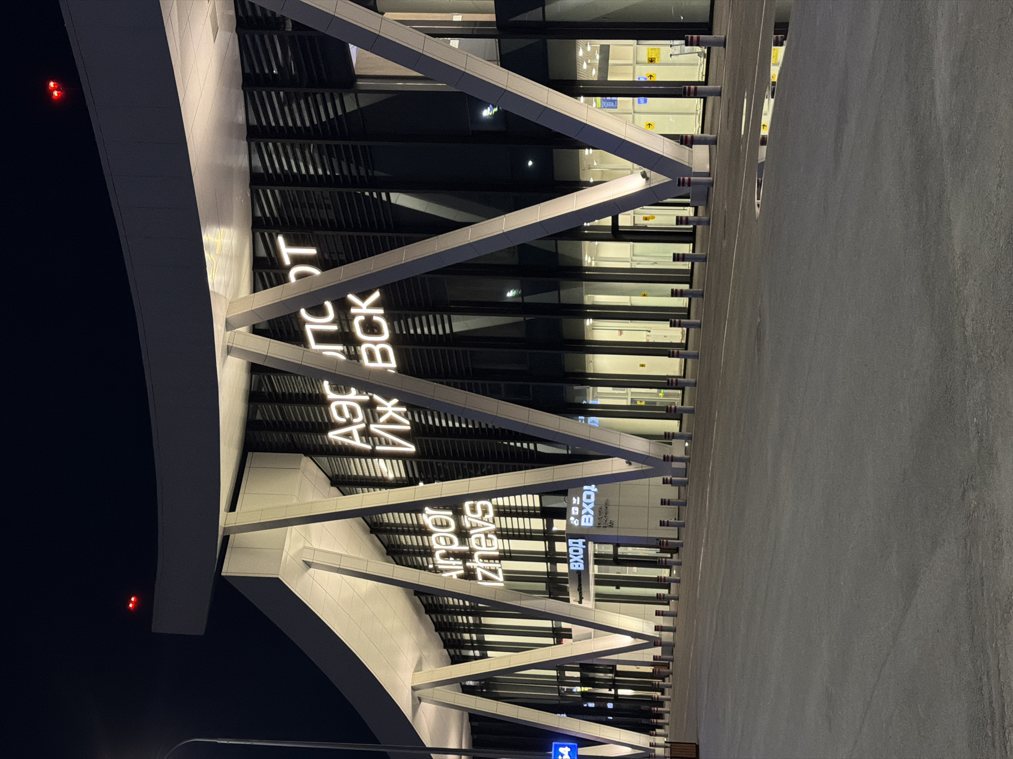 The image shows the modern facade of the Izhevsk airport at night. The building features a distinctive architectural design with large, angled white beams supporting a curved roof. The facade is primarily glass, reflecting the lights and the dark sky. Neon signs are visible, with "Airport Izhevsk" in English and what appears to be Russian text above it, possibly "Аэропорт Ижевск". Red aviation warning lights are positioned on the top edge of the roof. The area in front of the airport is a paved expanse, with rows of bollards lining the walkway and building entrance. Puddles of water are visible on the pavement, reflecting the lights. The overall scene conveys a sense of a contemporary, well-lit transportation hub operating at night.