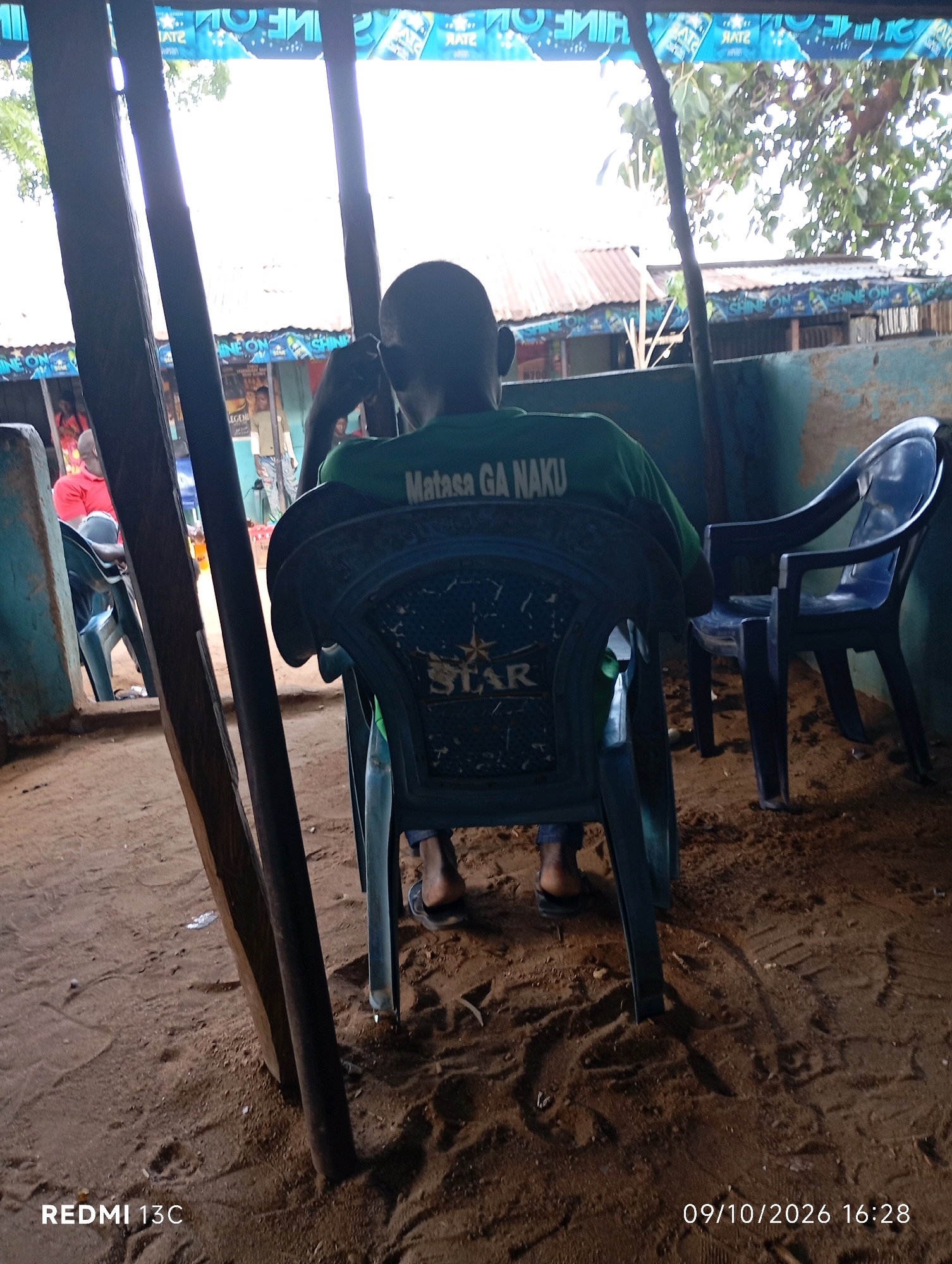 In Shendam, Nigeria, a person wearing a green shirt with the text "Matasa Ga Naku" is seated on a blue plastic chair, facing away from the camera. The person appears to be looking towards a stall or building in the background which has a corrugated metal roof and is decorated with banners that have blue and white lettering. The stall is lined with merchandise, including what appear to be bottles. Behind the person in the green shirt, another blue plastic chair is visible. The ground is covered in sand and dust, with some scattered leaves and debris. The scene suggests an outdoor setting, possibly a market or a roadside stall. The lighting indicates it is daytime. The overall atmosphere appears casual and informal.
