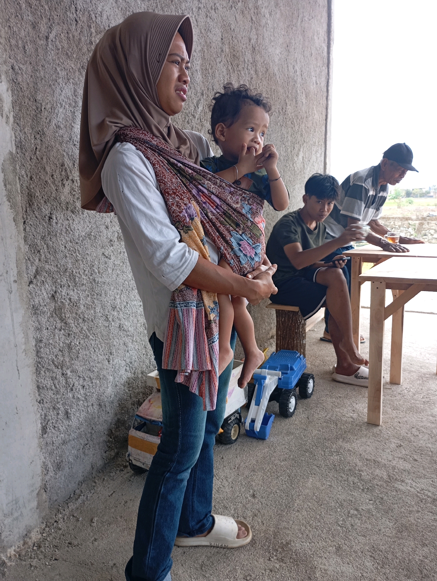 In Pacitan, Indonesia, a woman wearing a brown hijab and a white shirt is holding a young child wrapped in a patterned fabric sling. The child is looking off to the side with their fingers near their mouth. To the right, two men are seated at a wooden table. One man, wearing a dark green t-shirt, is looking towards the camera, and the other man, wearing a striped shirt and a black cap, is looking down at the table. There are two glasses of tea on the table. In the foreground, there are toy trucks on the concrete ground. The background appears to be an unfinished concrete wall and a cloudy sky.