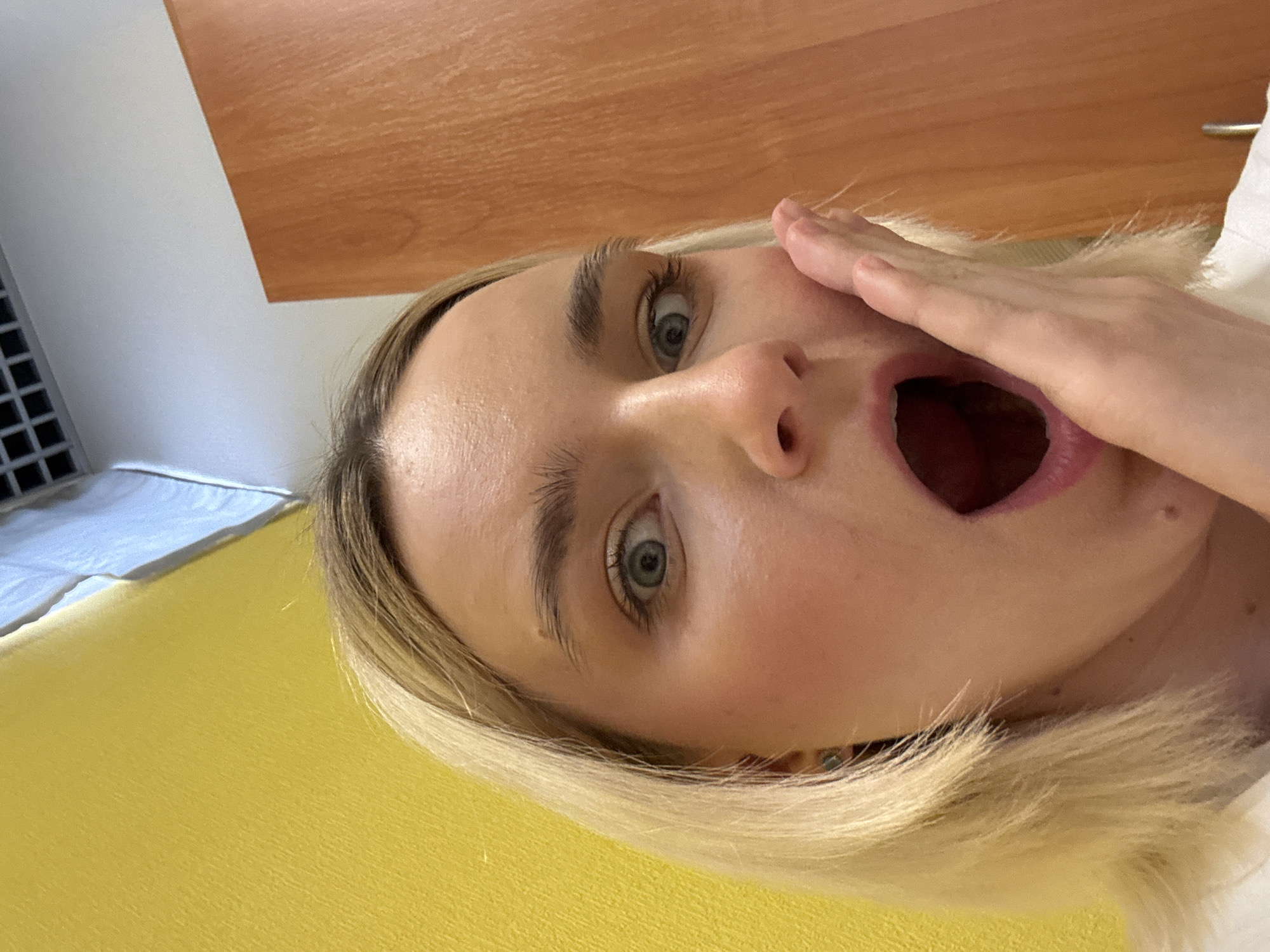 A close-up, rotated selfie shows a young woman with light blonde hair and fair skin. Her eyes are wide open and she has an expression of surprise or shock. Her mouth is agape, and her hand is partially covering her face. The background includes a wooden-looking surface and a yellow surface. The location is not discernible as being in Saint Petersburg, Russia, or any other specific location.