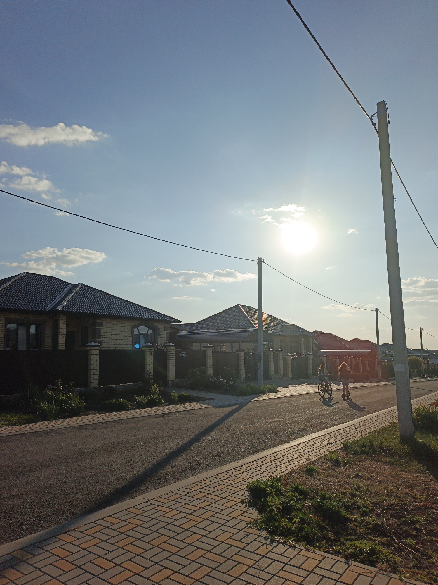 This is a street scene in Repnoe, Russia, on a sunny day with a bright sun and scattered clouds. The street is paved with asphalt, and a sidewalk of interlocking paving stones runs along it. On the left side of the street are modern houses with dark tiled roofs and brick and stucco exteriors, separated by dark fences. On the right side of the street, further down, are more houses with red tiled roofs. Two children are visible on the street: one is riding a bicycle, and the other is on a scooter. Both are moving away from the camera. Telephone poles and wires are present, casting long shadows across the scene. The overall impression is of a quiet suburban neighborhood in the late afternoon.