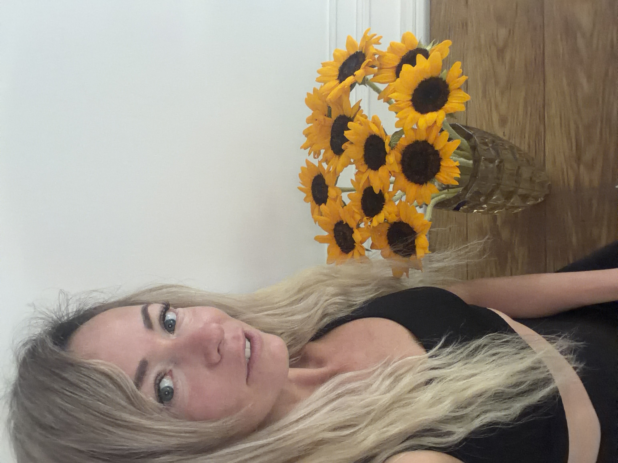 A woman with blonde hair is in the foreground, looking to her left. She is wearing a black athletic top. To her right is a bouquet of sunflowers in a glass vase. The sunflowers are bright yellow with dark centers. The background is a plain white wall. The scene appears to be indoors, likely in a home. There is no visible indication of the location in Xuân Trường - Đà Lạt, Vietnam.