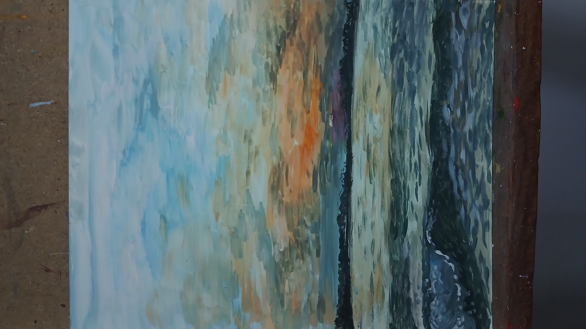 The image shows an oil painting of a seascape at sunset. The sky is filled with brushstrokes of blue, orange, and gray, suggesting dramatic clouds. A dark silhouette of a treeline or landmass forms the horizon. The water in the foreground is rendered with various shades of green and blue, with white highlights indicating waves and foam. The painting is displayed on a wooden easel or surface. There are no people or specific landmarks visible that would indicate a location in Oral, Kazakhstan.
