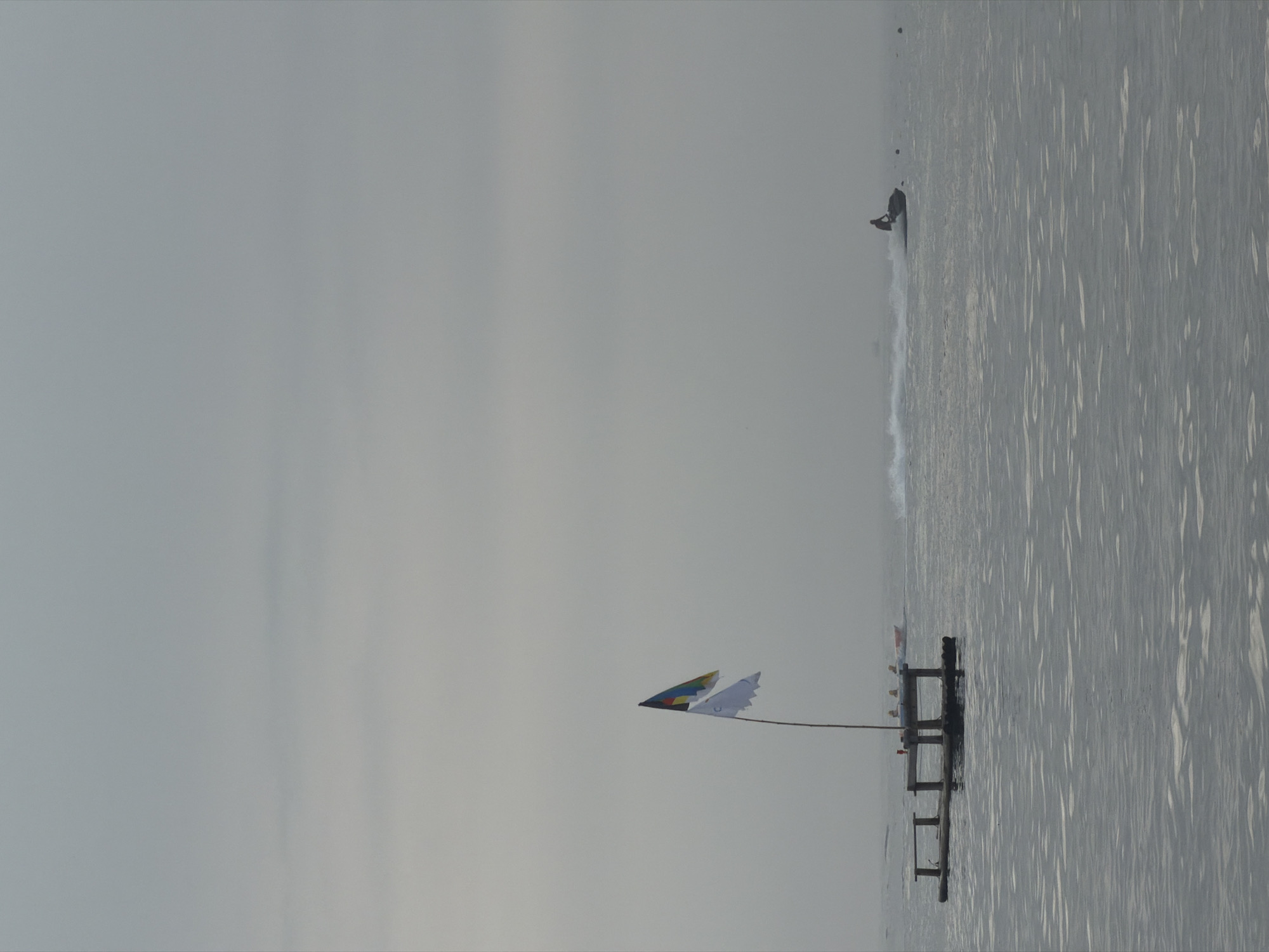 The image captures a serene, overcast day at sea, likely off the coast of Jeju, South Korea. The water is calm with gentle ripples, reflecting the muted, gray sky. In the foreground, a small, wooden platform or raft floats on the water, supported by submerged logs. A tall pole rises from the center of the platform, adorned with two flags. The upper flag is triangular with sections of yellow, blue, and a darker color, possibly black or dark green, while the lower flag is white with ragged edges.
Further out in the water, a jet ski speeds across the surface, leaving a white spray in its wake. A single person is visible on the jet ski, silhouetted against the hazy horizon. To the left of the jet ski, a small, orange boat with several figures on board is moving, also creating a slight disturbance on the water. The overall atmosphere is tranquil, with the vastness of the sea and sky dominating the scene, broken only by the activity of the watercraft.