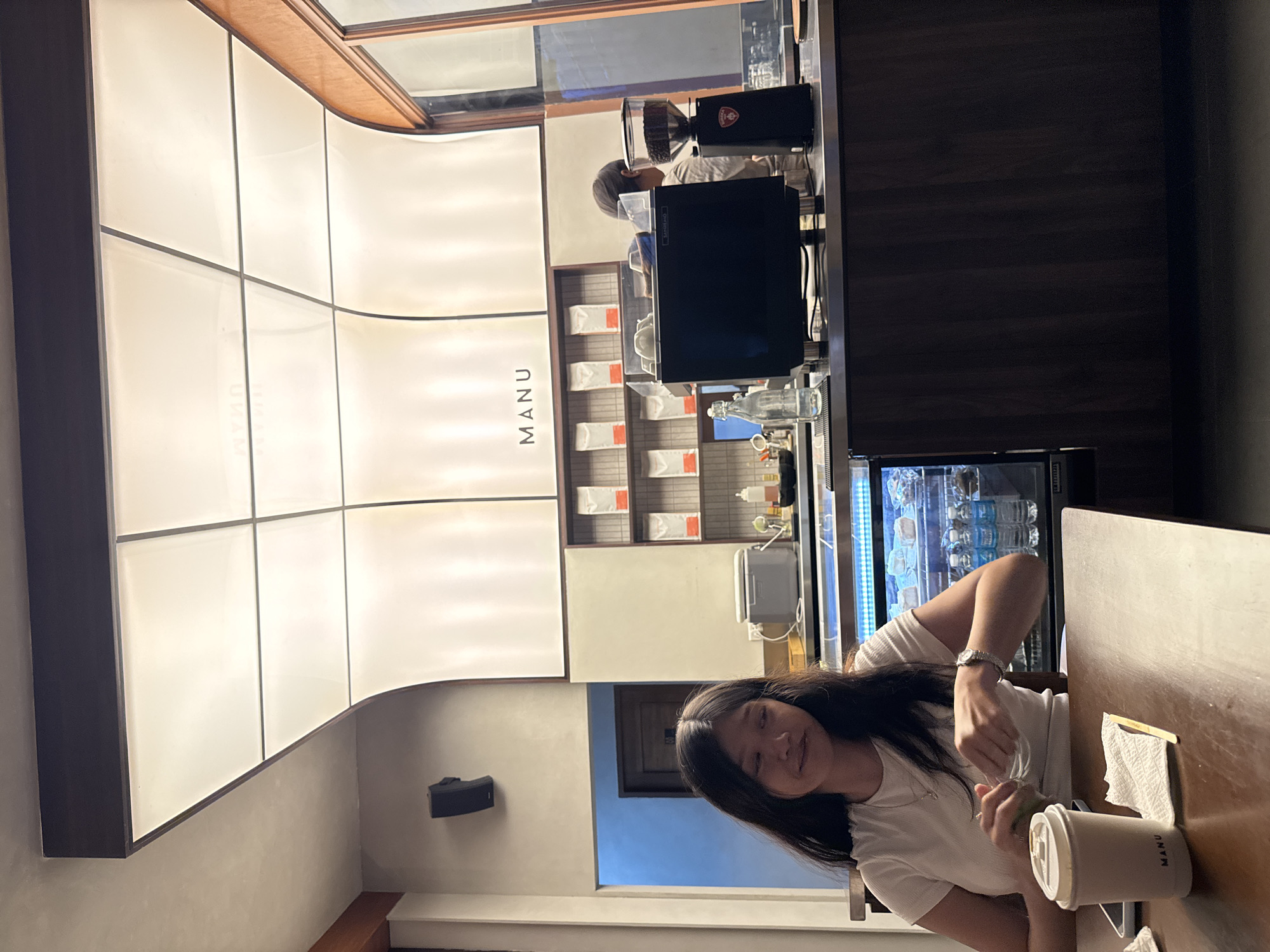 A young woman sits at a table in the coffee shop Manu in Davao City, Philippines, stirring a beverage in a paper cup. The cafe has a contemporary design with white panel walls with dark wood trim, and a retail display of coffee beans behind the counter. A barista is visible behind the counter, and a refrigerated display case stands next to the table.