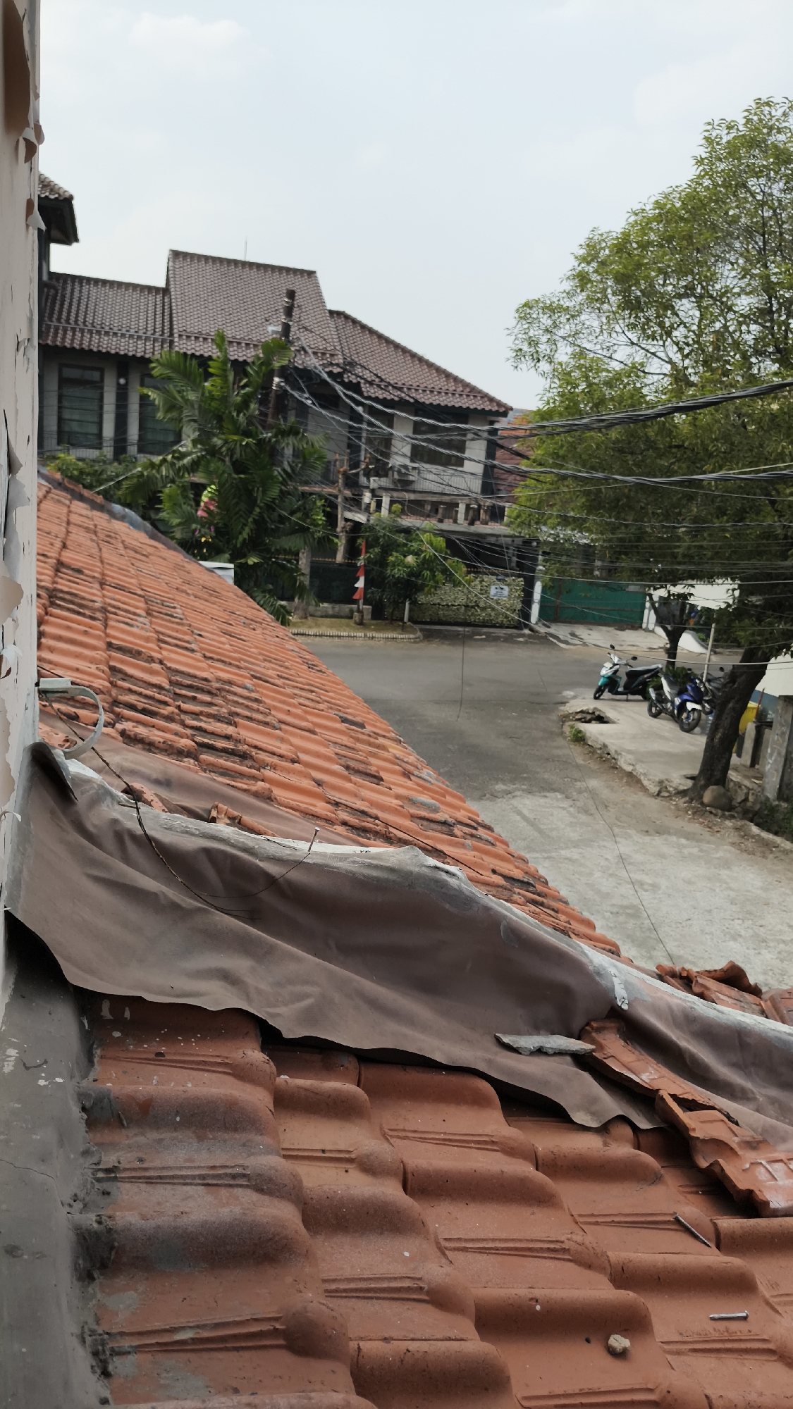 The image depicts a view looking down at a tiled roof, with a street and buildings in the background. The roof is made of reddish-orange tiles, some of which appear weathered or damaged. A dark brown tarp is draped over a section of the roof. In the background, there is a street with a few motorcycles parked on the side. The buildings in the background are multi-story structures with tiled roofs, characteristic of residential areas. Trees and power lines are also visible. The overall scene suggests an outdoor environment in an urban setting. It is likely located in a residential neighborhood of South Jakarta, Indonesia, given the architectural style of the buildings and the presence of motorcycles. The sky is overcast, indicating a cloudy day.