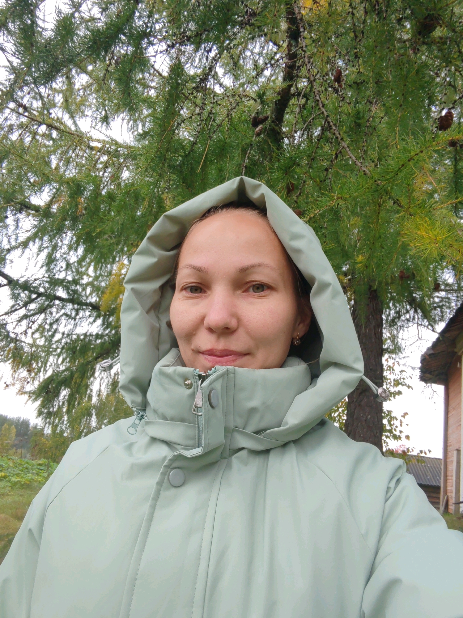 A woman stands outdoors, facing the camera, with a light green hooded jacket zipped up. She has light brown hair pulled back and is wearing a small, star-shaped earring in her left ear. The background features a large evergreen tree with pinecones and some yellowing leaves, suggesting an autumn setting. To the right, a portion of a building with a dark roof and brick siding is visible. In the distance, a hint of foliage and a body of water can be seen. The woman appears to be in a rural or semi-rural area.