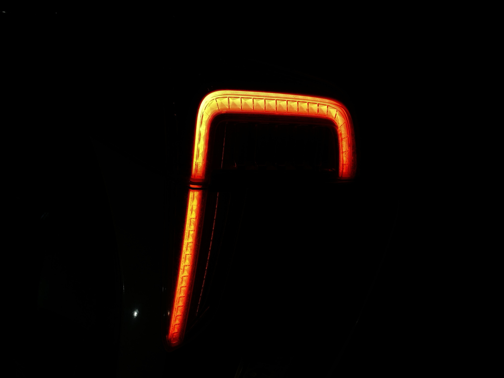 The image displays a close-up of a bright, glowing red-orange light against an entirely black background. The light forms a distinct angular shape, resembling the upper-left portion of a bracket or an inverted "L" (┐). It consists of a horizontal segment across the top and a vertical segment extending downwards on the left side.
This illuminated element appears to be a modern vehicle's taillight or brake light, characterized by its intense glow and structured design. Along the inner edge of the glowing light, there is a visible texture composed of numerous small, raised rectangular or square elements, suggesting an intricate internal design within the light fixture. The light emits a vivid, consistent red-orange hue.
The surrounding environment is pitch black, indicating that the photograph was taken at night or in an extremely dark setting. There are no other discernible objects, people, or landscape features visible in the darkness, placing sole emphasis on the luminescent subject. A very faint, small white dot, possibly a distant reflection or a minor light source, can be observed in the lower-left area of the dark background, but it is unobtrusive. No specific activity or event is taking place other than the light being illuminated. There is no visible text.