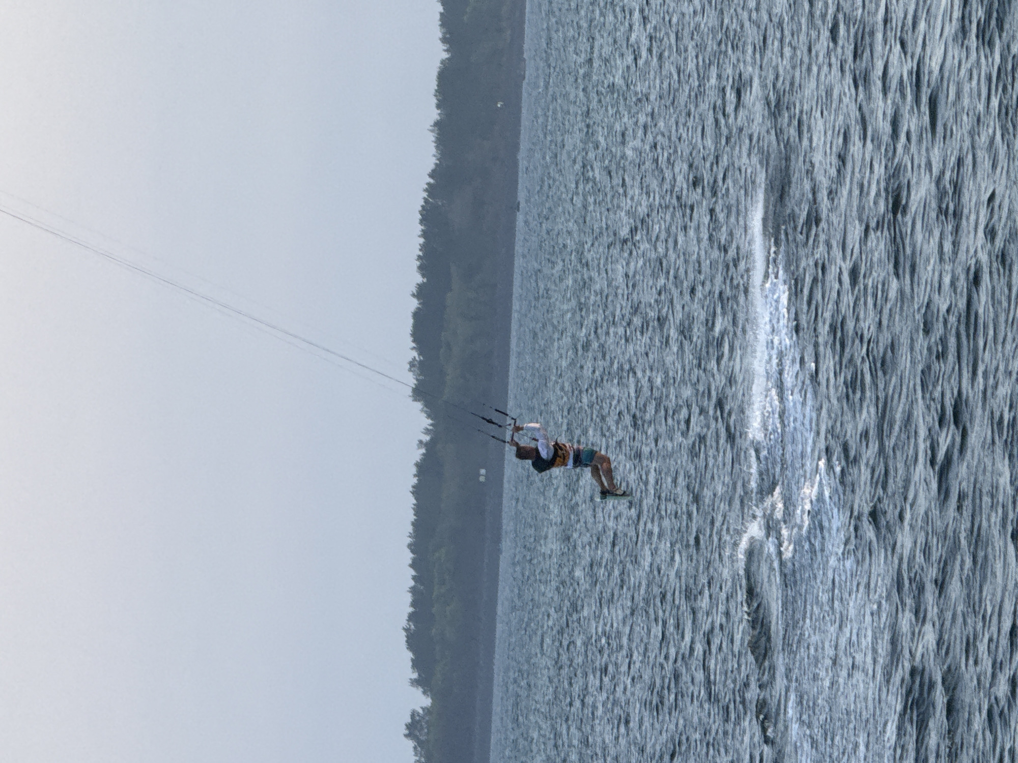 A single person is kitesurfing on a choppy, dark blue body of water. The kitesurfer is airborne, with their body angled slightly backward. They are wearing a life vest over a long-sleeved white shirt and patterned shorts, with a green board strapped to their feet. The kitesurfer is holding onto a control bar connected to lines that extend upwards out of the frame. In the background, a dense, dark green tree line forms a horizon against a pale, overcast sky. The water exhibits small waves and white spray near the kitesurfer, indicating movement. The scene appears to be outdoors, likely on a large body of water, potentially a bay or ocean.