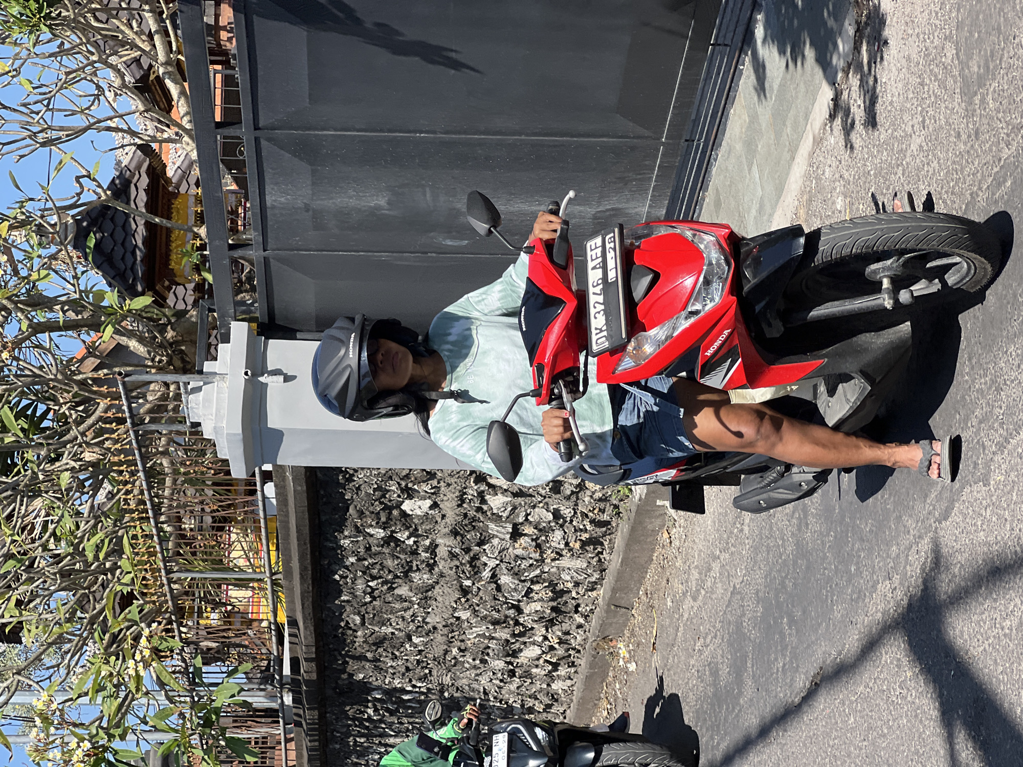 A person wearing a light blue tie-dye long-sleeved shirt, denim shorts, flip-flops, and a black helmet is riding a red Honda scooter in Denpasar, Indonesia. The scooter's license plate reads "DK 3246 AFF". Another person is riding a black scooter in the background to the left, wearing a green jacket. The scooters are on a paved road next to a textured gray stone wall. Behind the wall, there is a black metal gate and a building with a tiled roof and decorative elements, partially obscured by trees with white flowers. The sky is clear and blue.