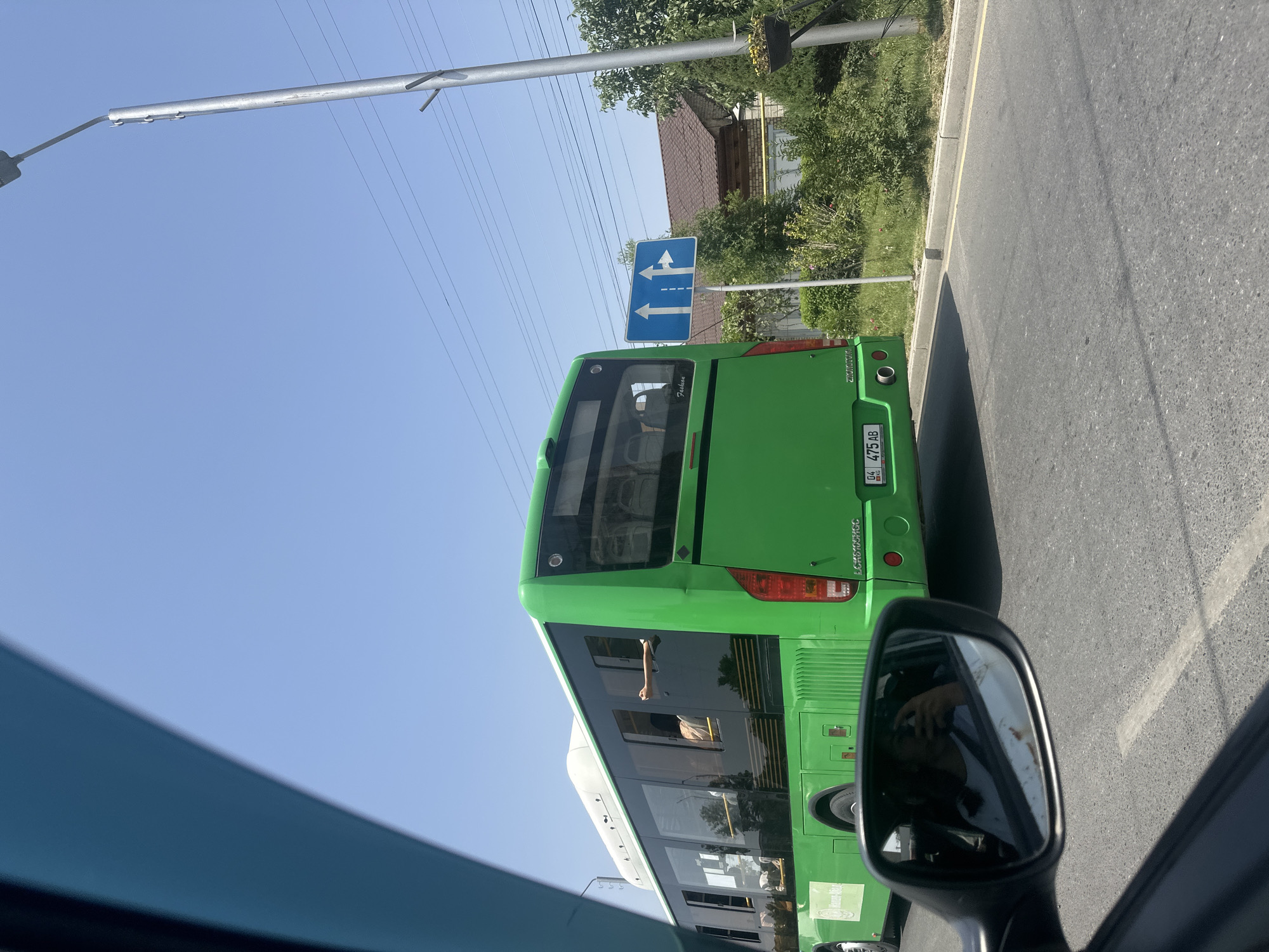 An outdoor, eye-level, medium shot captures a bright green bus from its rear quarter in Manas, Kyrgyzstan. The bus, identified by the license plate "04 475 AB KG", is moving on a paved road. A white building with a tiled roof and green trees are visible in the background, with a street lamp to the right. The sky is clear and blue. Inside the bus, through its windows, a person can be seen seated. A car's side mirror on the left reflects the driver taking a photo. The scene appears to be a sunny day.