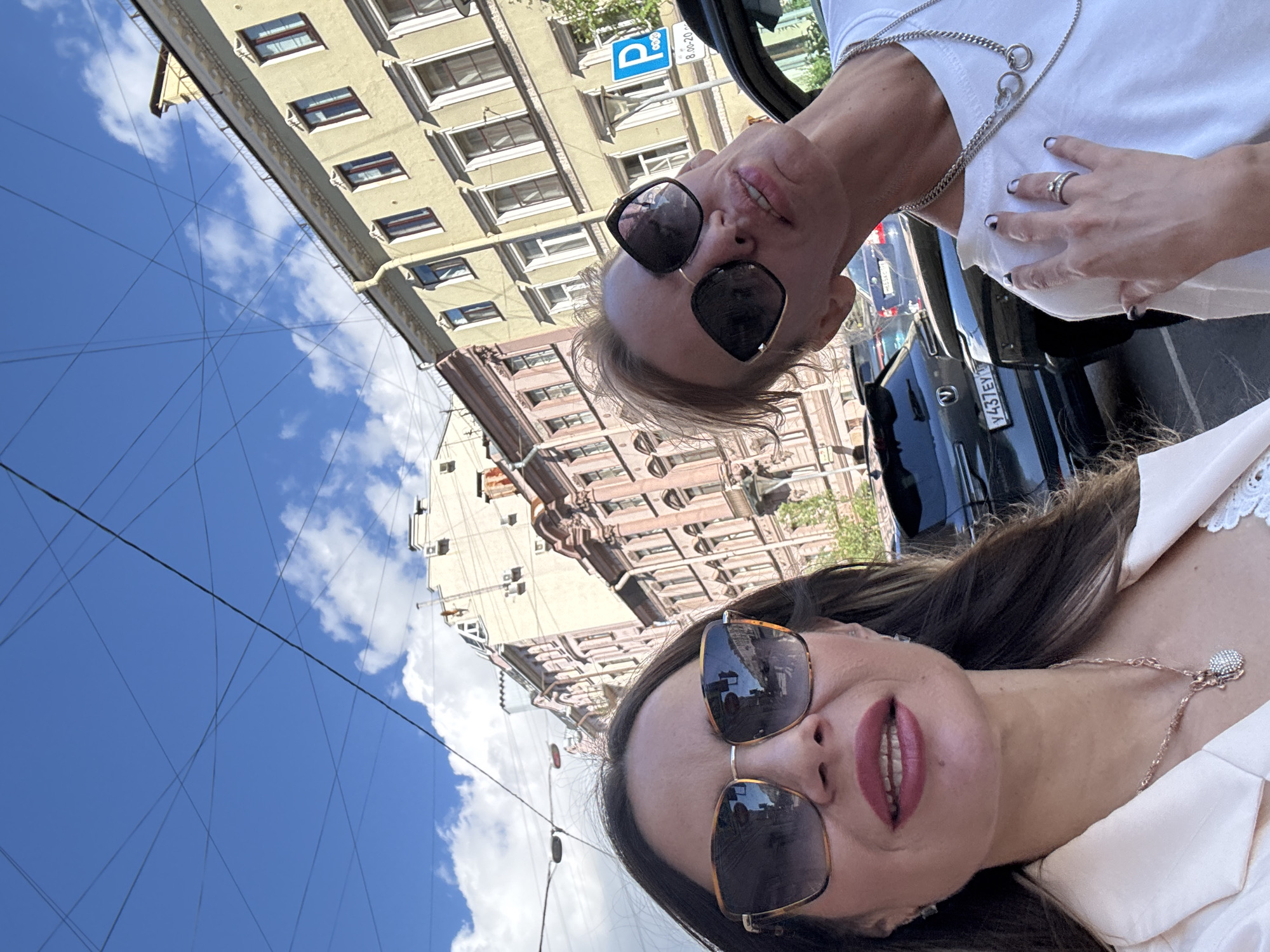 Two women are taking a selfie outdoors on a street in Saint Petersburg, Russia. The woman on the left is wearing sunglasses and a light-colored jacket. She has dark hair and is smiling. The woman on the right is also wearing sunglasses and a white t-shirt. She has blonde hair pulled back and is looking up at the camera. They are standing in front of a row of buildings with visible architectural details. Overhead, numerous electrical wires crisscross against a blue sky with scattered clouds. In the background, cars are parked along the street, and a blue sign with a white "P" indicating parking is visible. The overall setting suggests a sunny day in an urban environment.