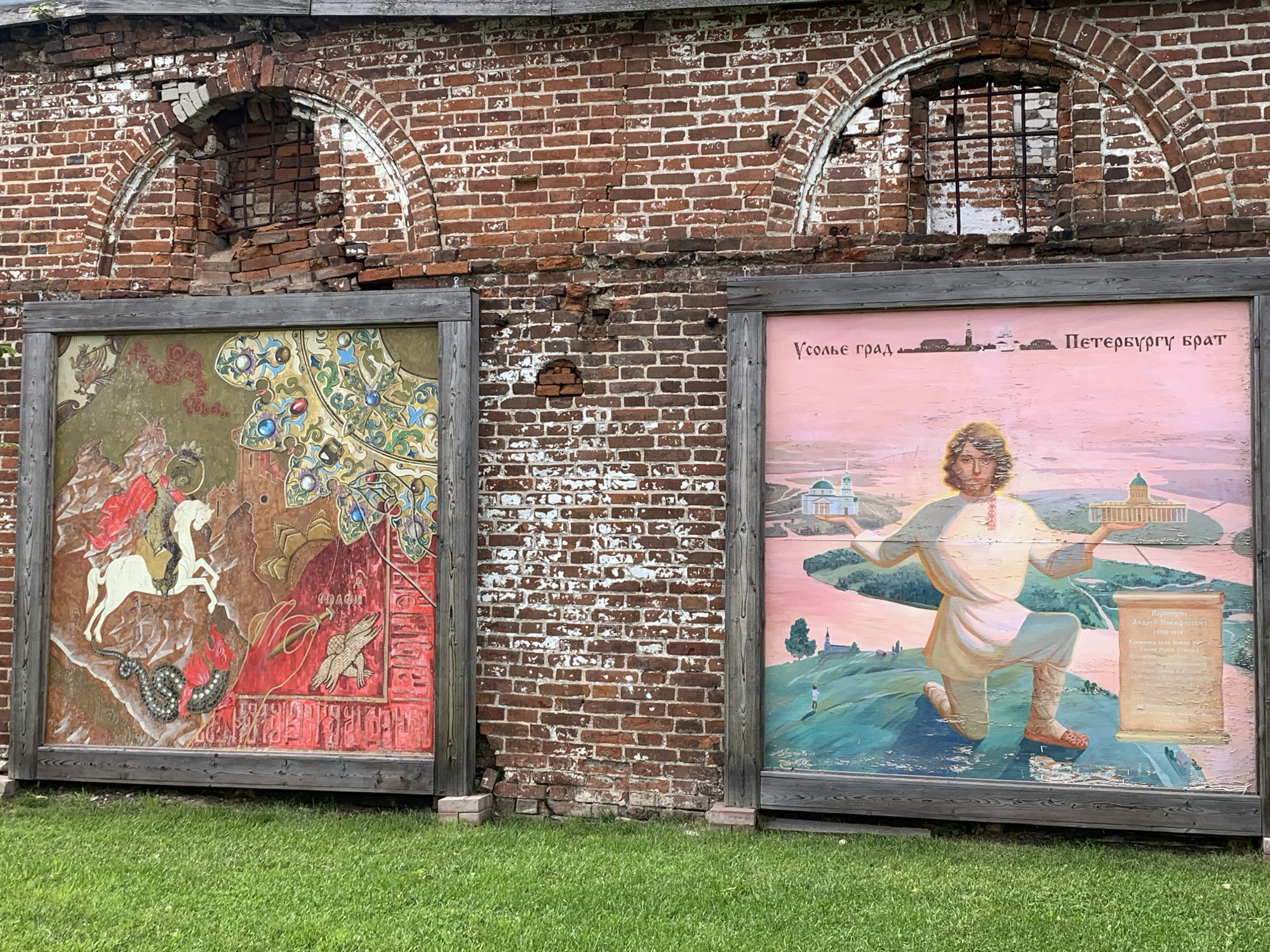 The image shows a section of a weathered brick wall in the city of Usolye, Russia. Two large, framed paintings are displayed in front of the wall. The painting on the left depicts a religious scene with a figure on a white horse fighting a dragon, rendered in rich, vibrant colors with intricate patterns and gold accents. The painting on the right features a landscape with a kneeling figure holding architectural models in each hand, set against a pink sky with buildings and trees. Text in Cyrillic script is visible on both paintings, suggesting they are historical or commemorative in nature. The wall itself is in a state of disrepair, with crumbling bricks and arched window openings covered by metal grates. The foreground is covered with green grass. The overall scene suggests an outdoor exhibition or historical site in Usolye.