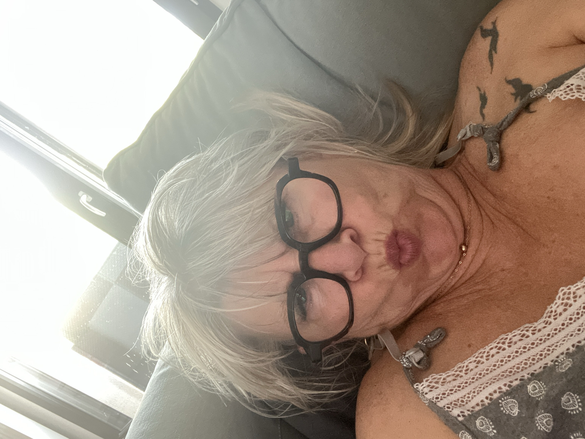 This is a selfie of a woman with blonde hair and glasses, lying on a couch. The woman is wearing a grey and white patterned top with lace trim. She has tattoos on her upper arm and shoulder. Her lips are pursed, and she is looking directly at the camera. The lighting suggests it is daytime, with sunlight streaming in from a window, creating a bright, possibly hazy effect. The couch appears to be made of a dark grey fabric. There is no visible text in the image. The setting appears to be indoors, likely a living room.