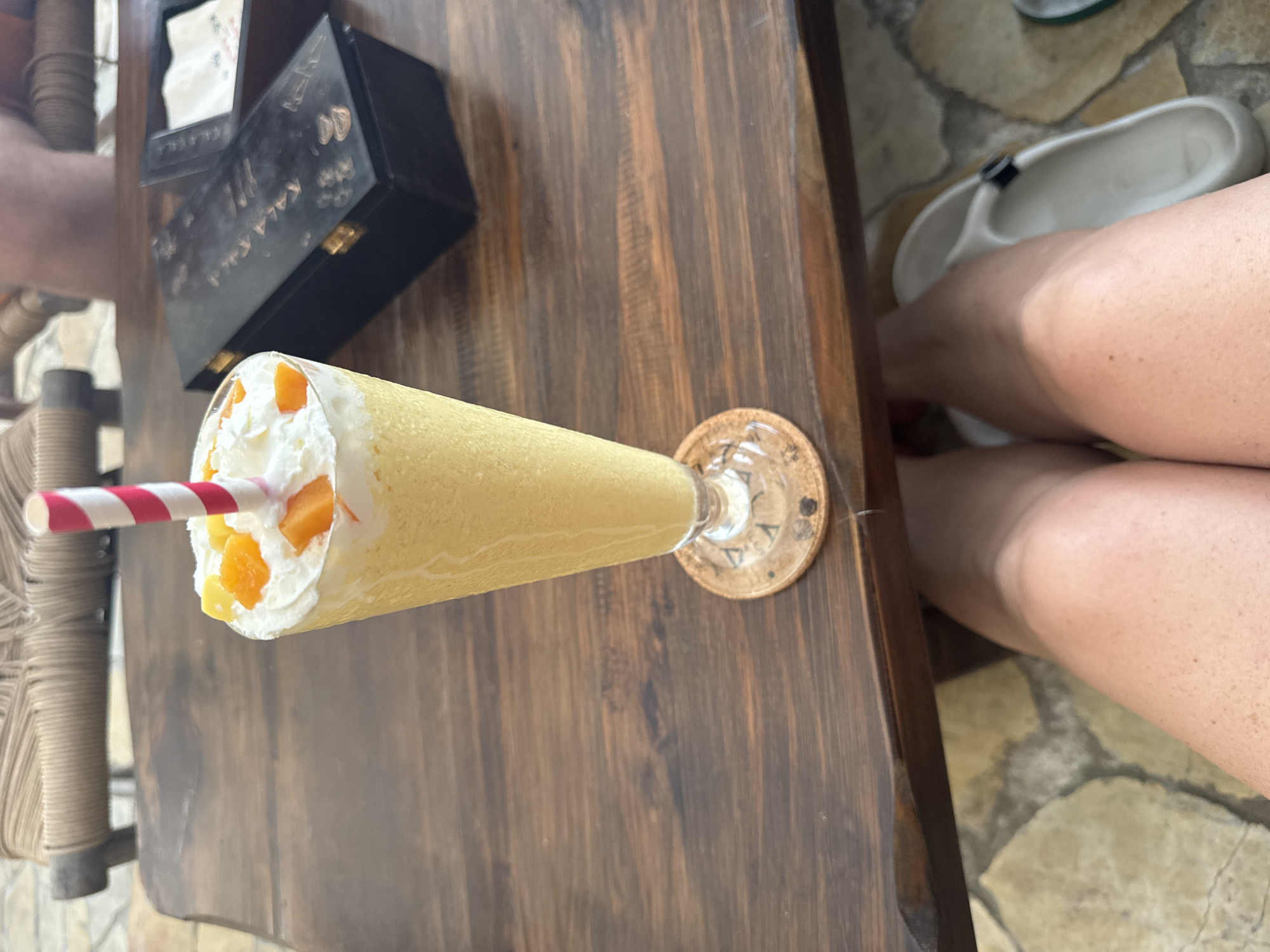 A mango smoothie is served in a tall glass on a wooden table. The smoothie is topped with whipped cream and mango chunks, and a red and white striped straw is visible. To the left of the smoothie, there is a black box. To the right of the table, a person's legs are visible, with one foot wearing a white sandal. The background shows a stone-paved area. The location is likely a café or restaurant in Đà Nẵng, Vietnam.