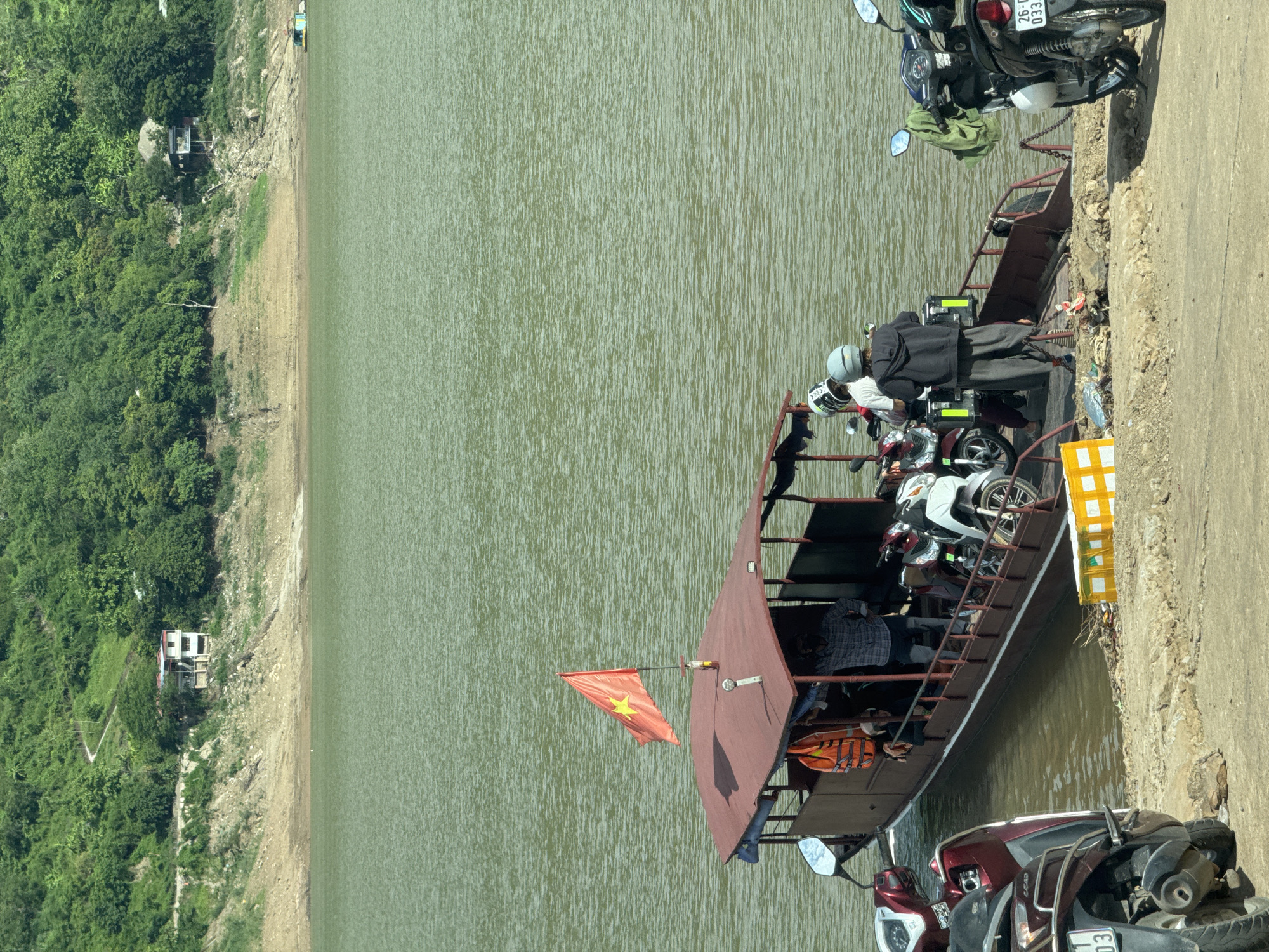 This image depicts a ferry boat docked at a riverbank in Tân Phong, Vietnam. The ferry, a long, flat-bottomed vessel with a red canopy and a Vietnamese flag, is laden with several motorbikes and passengers. Two individuals are visible on the ferry, one standing near the rear and another seated. Two motorbikes are positioned on the ferry's deck, and two more are waiting to board on the riverbank. One motorbike on the riverbank is maroon, and the other is black. The river is a calm, green body of water. In the background, a lush green, tree-covered hillside rises steeply from the riverbank. Several small houses are scattered across the hillside, and a winding path is visible ascending the slope. The riverbank itself is a mixture of dirt and rocks. The overall scene captures a moment of transportation and daily life in a riverside community.