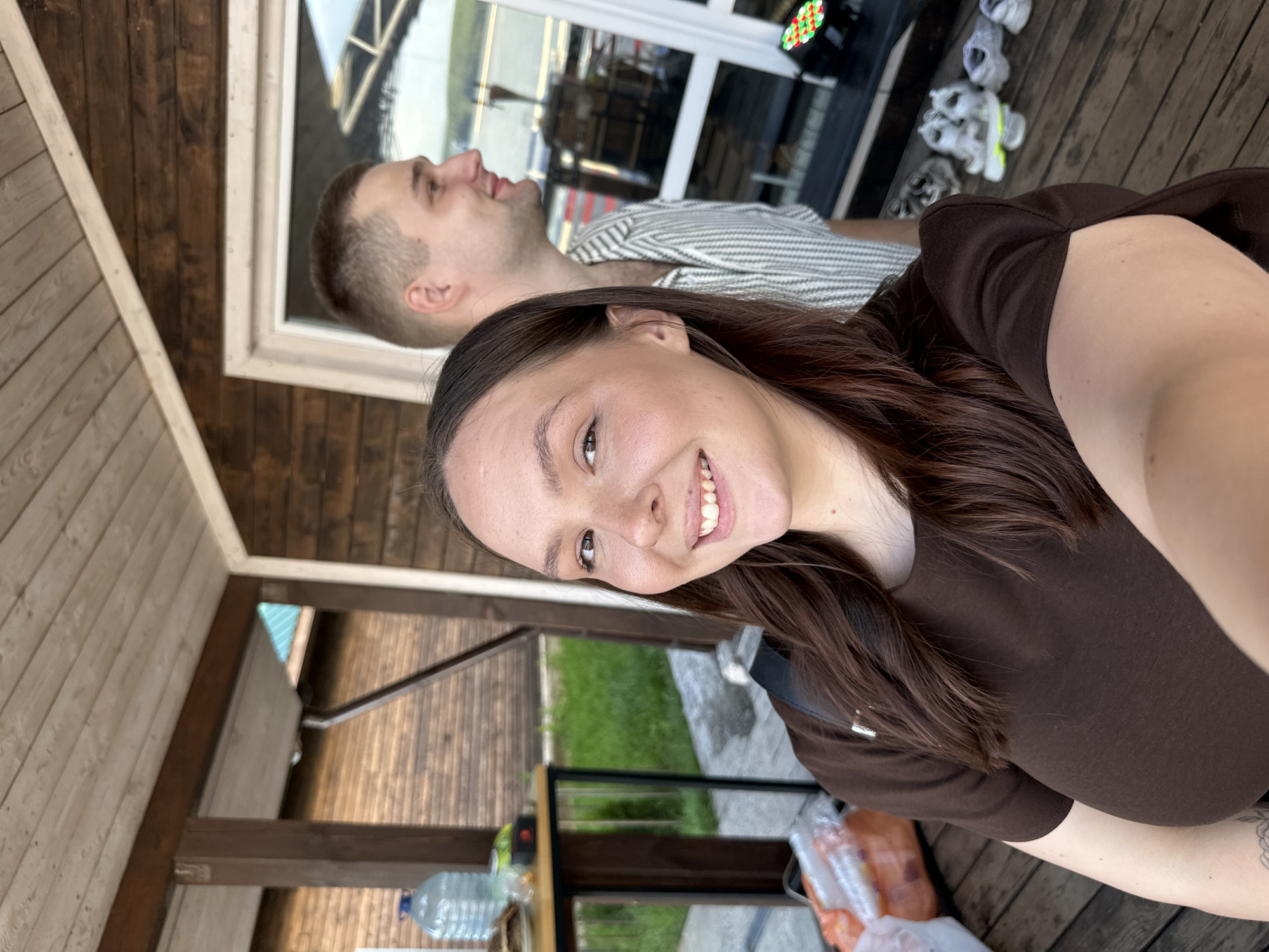 A woman and a man are standing outside a wooden building. The woman is in the foreground, smiling at the camera and holding it up for a selfie. She has long, dark brown hair and is wearing a brown t-shirt. Behind her, the man is looking to the side and smiling. He has short, light brown hair and is wearing a striped shirt. They are on a wooden deck, with a wooden building to their right. There are several bottles of water and some plastic bags on a table to the left. In the background, there is grass and more wooden structures. The scene appears to be a casual outdoor setting. It is not possible to identify the specific location within the city of Chaikovsky, Russia, based solely on the visual information provided.