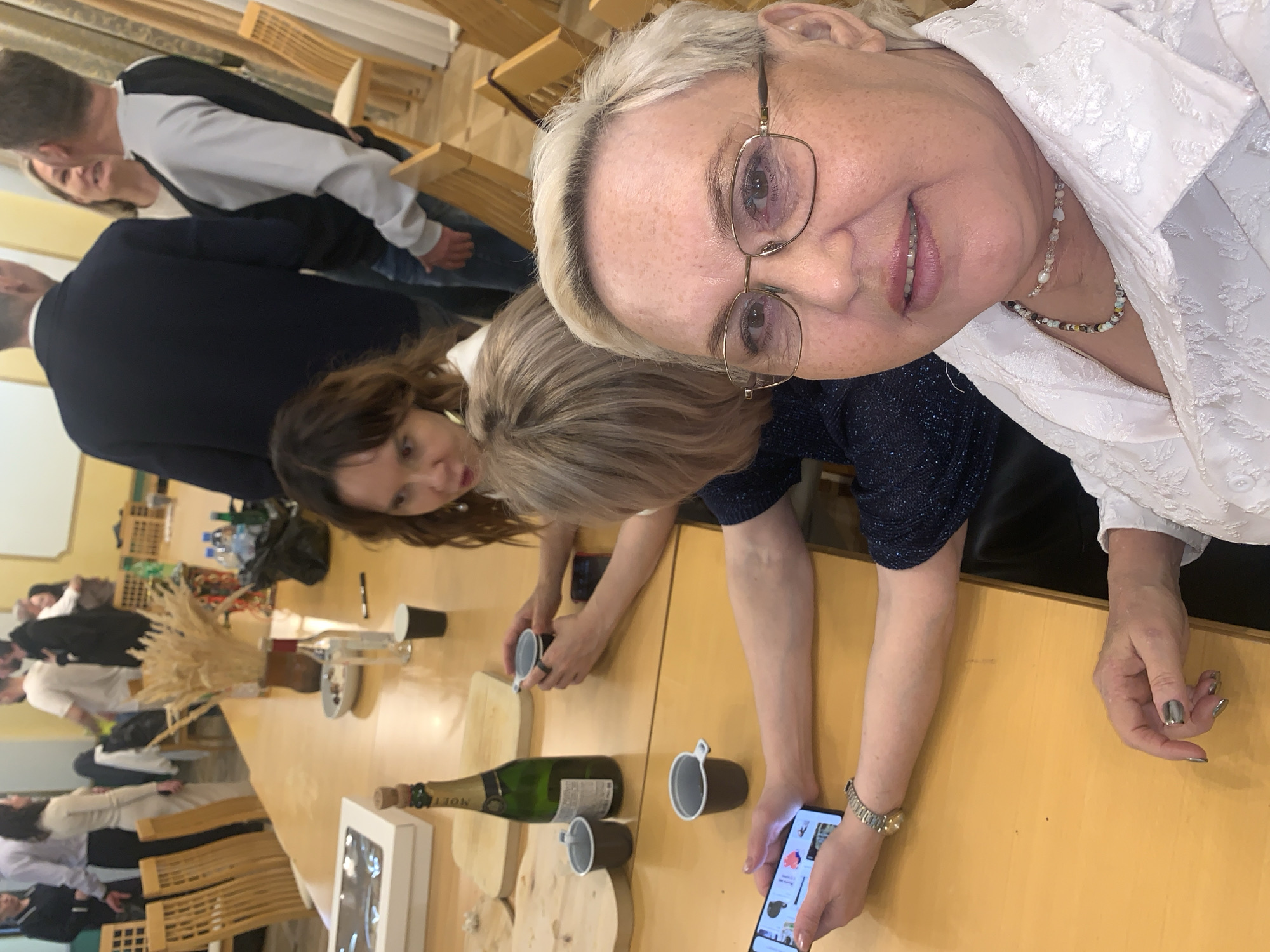 The image depicts a social gathering around a long wooden table. Present are several individuals, most notably a woman with blonde hair and glasses, positioned closest to the camera, seemingly taking a selfie with her mobile phone. She is wearing a white blouse and a necklace. Beside her is another woman with brown hair, sitting at the table.
On the table are various items, including a bottle of Moet & Chandon champagne, several dark-colored cups, a box, and what appears to be a decorative arrangement of dried wheat. Other people are situated at the table further away from the camera.
The setting seems to be indoors, likely a hall or a large dining room, characterized by chairs and tables with people seated.
I can infer the image was taken in Chaikovsky, Russia because that was stipulated in the prompt.