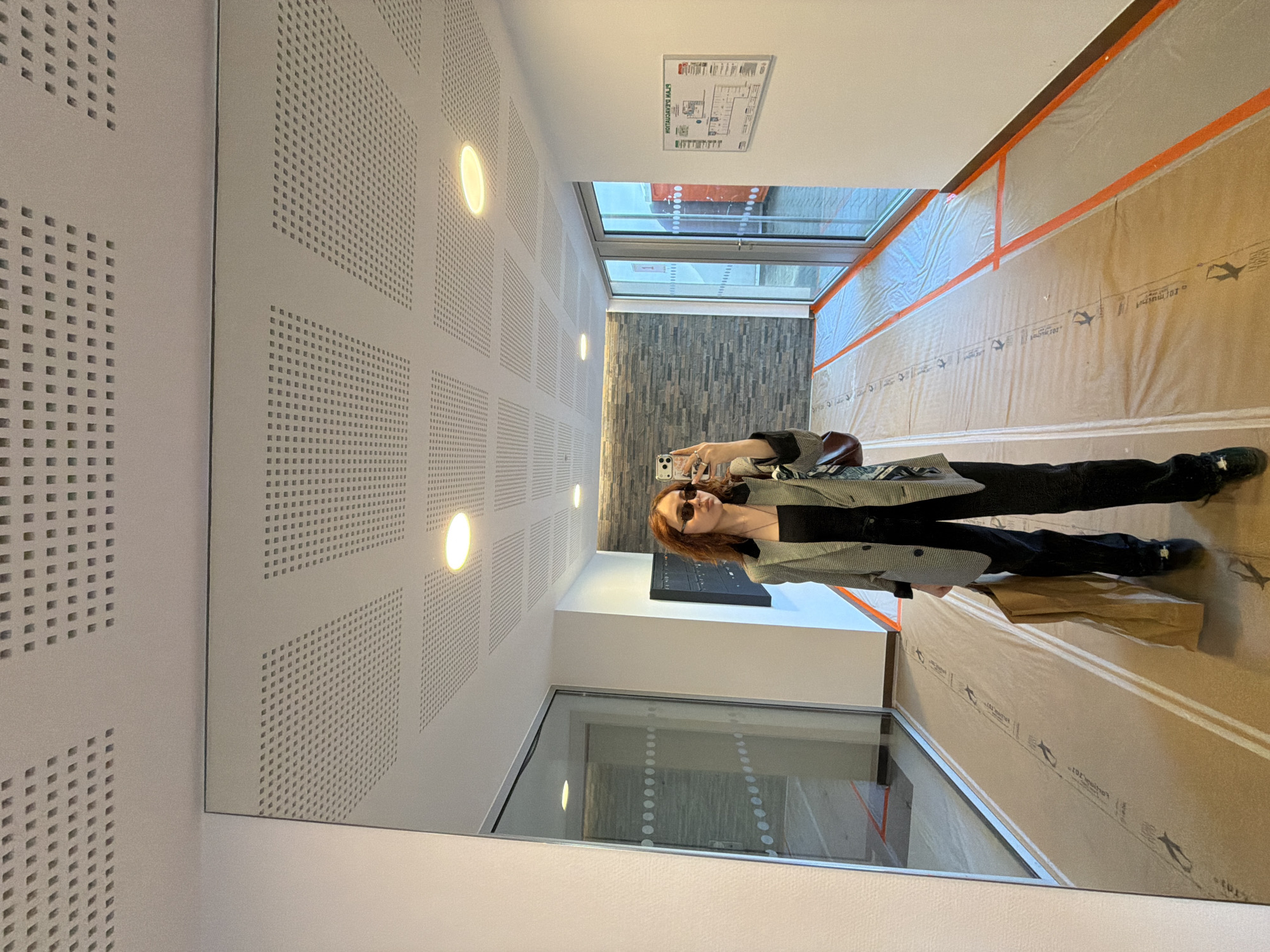 A person is captured in a mirror selfie within a brightly lit indoor corridor. The subject is a woman with reddish-brown hair and dark sunglasses, holding a smartphone with a decorated case to take the picture. She is dressed in a patterned blazer over a dark top, dark pants, and dark shoes. A brown paper bag and another bag rest on the floor beside her.
The corridor features a large mirror on the left wall, reflecting the woman and the opposite side of the hallway. The ceiling is equipped with square perforated acoustic panels and multiple circular recessed lights, which cast a warm glow. At the far end of the corridor, large windows offer a view of the outside, and a section of wall is covered in dark, textured, possibly stone-like panels.
The right wall of the corridor is covered in light brown protective sheeting, secured with orange adhesive tape. This sheeting has repeated text and a stylized bird or airplane logo, with visible text including "® Formwork". A white sign, visible on the ceiling above the window area, is a "PLAN D'ÉVACUATION" (Evacuation Plan), appearing reversed in the mirror. The presence of the protective sheeting suggests the area might be undergoing renovation, construction, or recent completion. The lighting indicates it is likely daytime. The setting appears to be a modern building, possibly in an office or institutional context in Strasbourg, France.