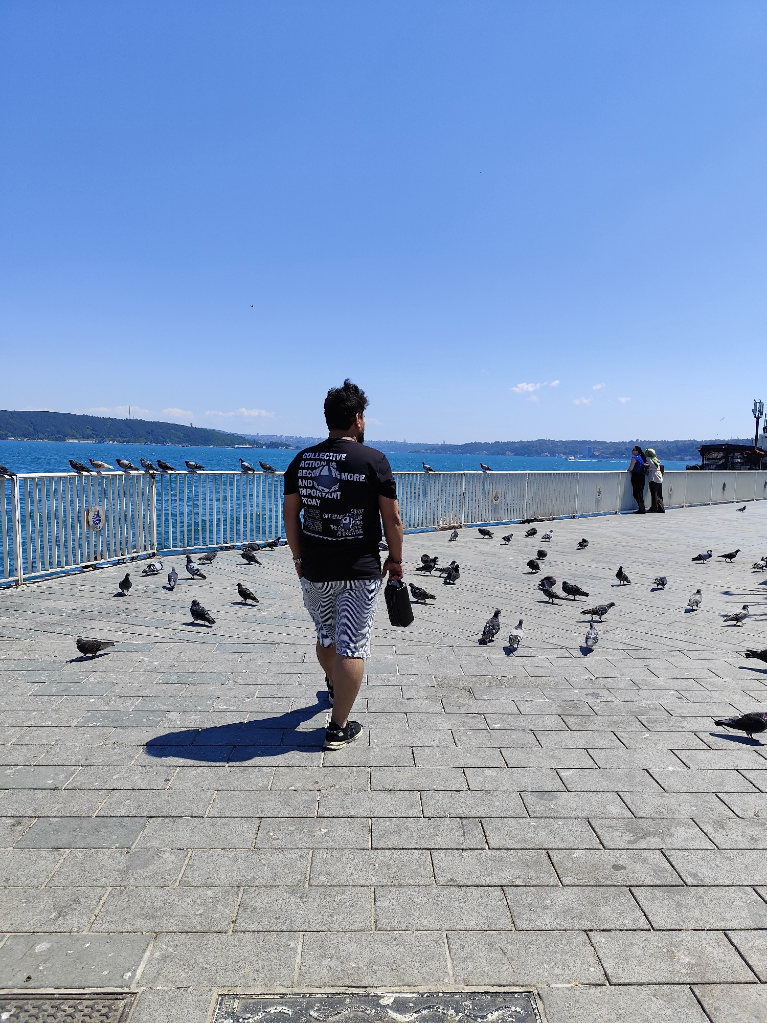 An overcast, daytime shot of a person walking on a stone-paved promenade next to a body of water. The person, a man with dark hair, is wearing a black t-shirt with text on it and striped shorts. He is carrying a dark bag in his right hand. Pigeons are scattered across the promenade. In the background, a white metal railing separates the promenade from the water. Beyond the water, a green, hilly landscape with buildings is visible under a clear blue sky. Two individuals are standing by the railing in the background, facing the water.