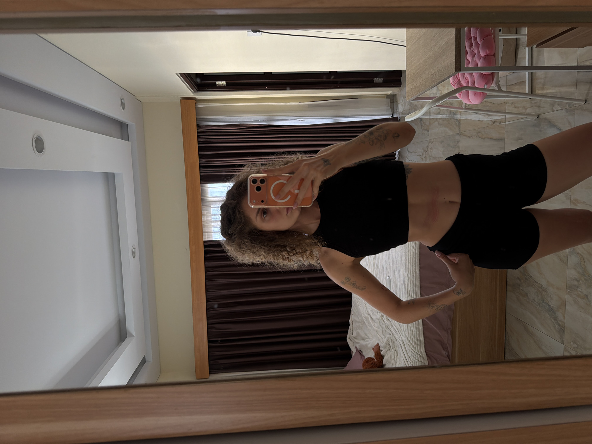 This image is a mirror selfie taken by a young woman in what appears to be a bedroom or private living space in North Kuta, Indonesia. The woman, with curly, light brown hair, is wearing a black crop top and black shorts. She holds an orange-cased smartphone, possibly an iPhone, to capture her reflection; a white MagSafe ring is visible on the phone case. She has several tattoos on her arms and subtle marks on her abdomen.
The large mirror, framed in light brown wood, reflects a bright and modern room. The walls are a mix of light cream/beige and white, with white recessed lighting fixtures on the ceiling. A prominent feature is a large window fitted with dark brown horizontal blinds and sheer white curtains, suggesting daytime. Below the window, a bed is visible with a light-colored, textured bedspread and a lavender-colored sheet. A small brown plush toy rests on the bed. To the right, a white metal stand holds a pink, soft object, possibly a cushion or folded fabric. The floor is covered in large, light-toned tiles with a subtle marbled pattern. A white electrical outlet and a thin black cable are visible on the cream wall.
The woman is the sole subject, engaged in the activity of taking a self-portrait. The scene conveys a relaxed, personal moment within a well-lit, contemporary indoor setting. No discernible text is visible besides the MagSafe design on the phone case.