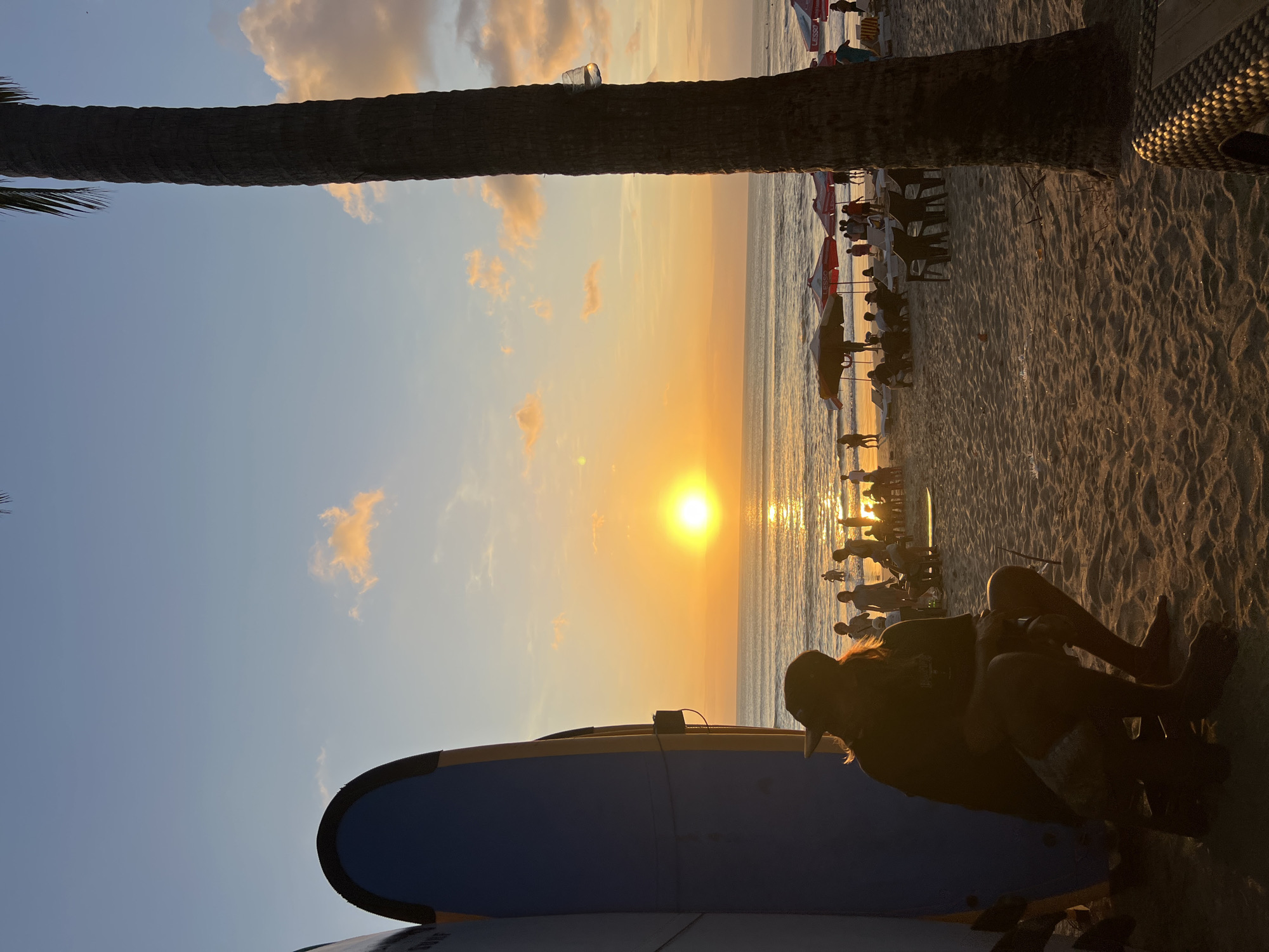 This image captures a sunset scene on Kuta Beach, Indonesia. The sun is low on the horizon, casting a warm, golden glow across the sky and the ocean. The water is calm, with gentle waves lapping at the shore.
In the foreground, a person is sitting on a low stool, silhouetted against the bright sky. They are wearing a hat and appear to be facing the ocean. To the left of the person, two surfboards are leaning against each other, their blue and yellow surfaces partially visible.
Further down the beach, several people are scattered around, some standing near the water, others seated at tables under umbrellas. Beach chairs and umbrellas are set up, indicating a recreational area. A tall palm tree stands prominently on the right side of the frame, its trunk dark against the colorful sky. A plastic cup is attached to the tree trunk.
The sand on the beach is textured with footprints and ripples from the receding tide. The overall atmosphere is peaceful and serene, characteristic of a beach at sunset.