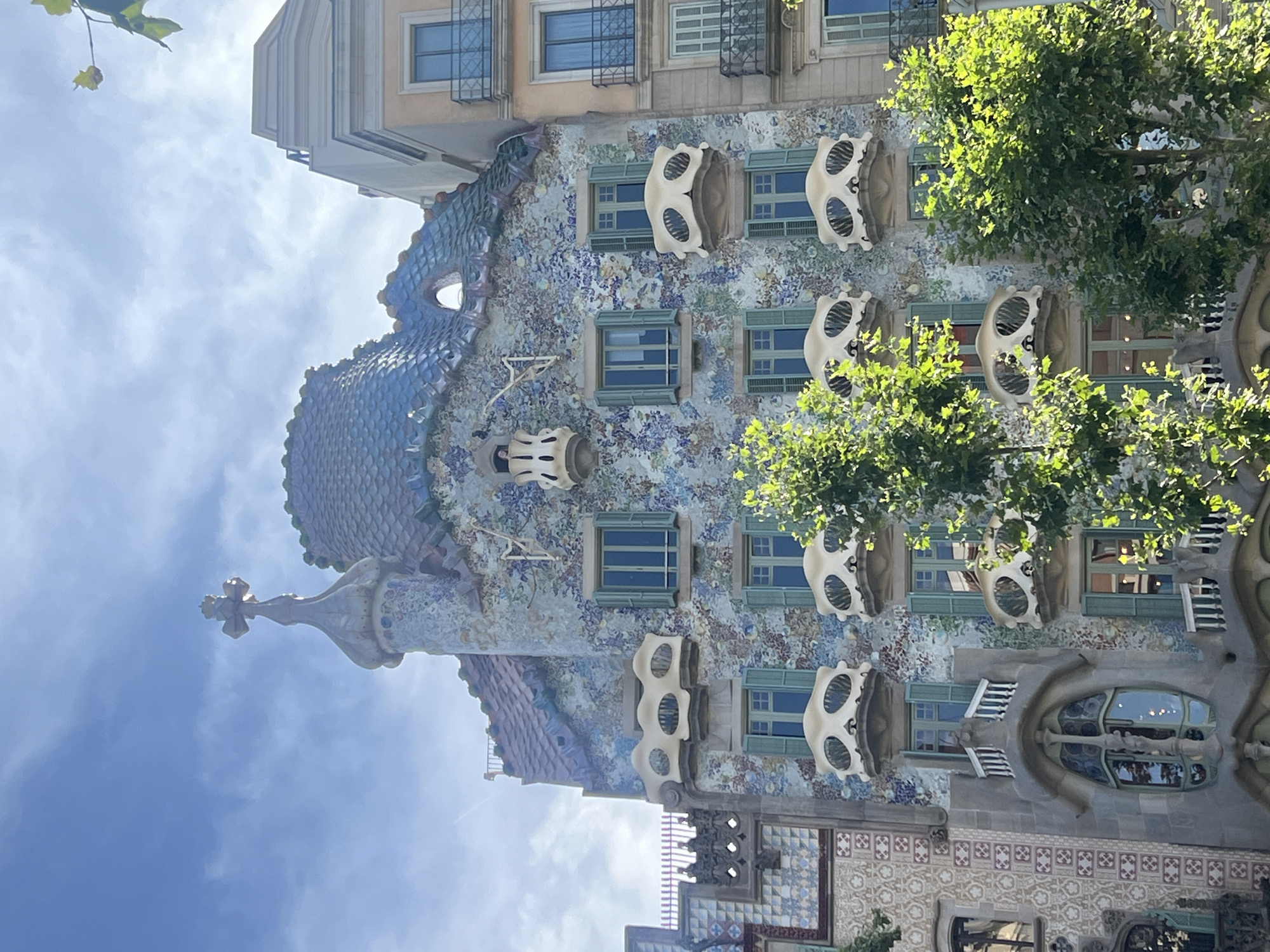 The image shows the exterior of Casa Batlló, a building located on Passeig de Gràcia in Barcelona, Spain. The building's facade is adorned with colorful mosaic tiles and features distinctive bone-shaped balconies. Several windows with green shutters are visible, and a tiled, dragon-like roof tops the structure. A cross tops a turret on the left side of the building. Trees with lush green foliage partially obscure the lower portion of the facade. The sky is blue with scattered clouds. Adjacent buildings, one lighter in color with balconies and windows, are visible on the right. No people are clearly discernible in the image.
