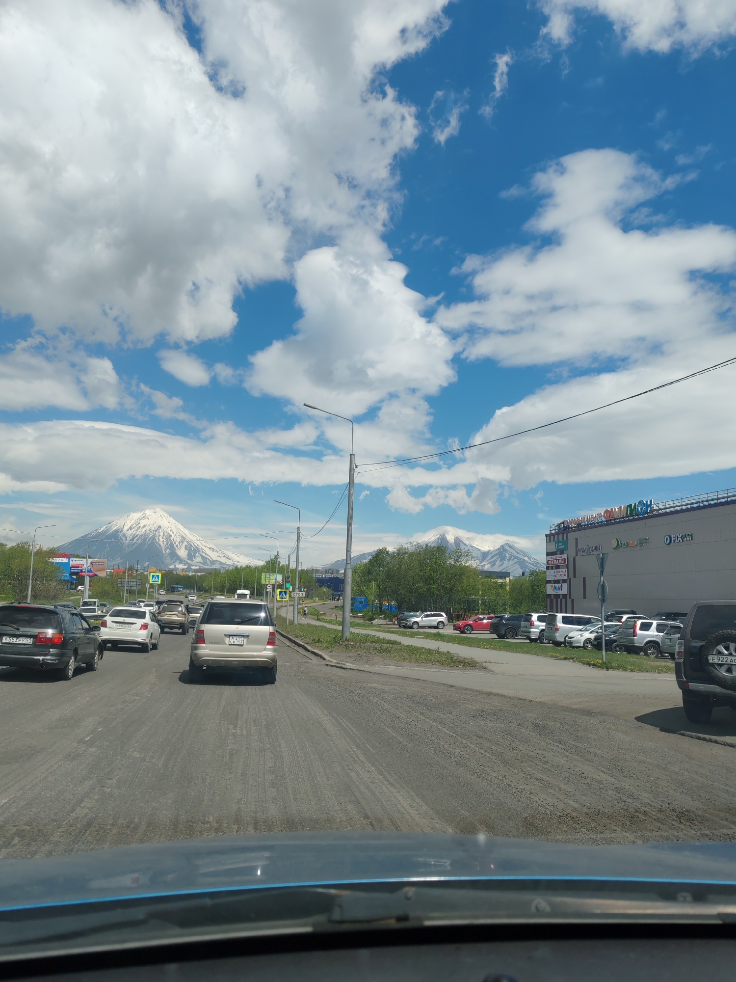 A wide-angle, eye-level shot captures a street scene in Petropavlovsk-Kamchatsky, Russia, on a partly cloudy day. In the foreground, the dashboard of a vehicle is visible, with the road ahead partially obscured by it. The road is paved with asphalt and appears somewhat worn, with visible tire tracks. A line of vehicles is traveling in the same direction as the viewer, including a black SUV in the left lane and a light-colored SUV in the right lane.
Further down the road, a large, snow-capped volcano dominates the background, its peak reaching into a bright blue sky dotted with fluffy white clouds. To the right of the volcano, a modern, multi-story building with a prominent sign displaying Cyrillic lettering is visible, likely a shopping center or commercial complex. Green trees line the sides of the road, and streetlights and traffic signals are present. Pedestrians can be seen walking on the sidewalk to the right. The overall impression is of a typical urban environment with a dramatic natural backdrop.