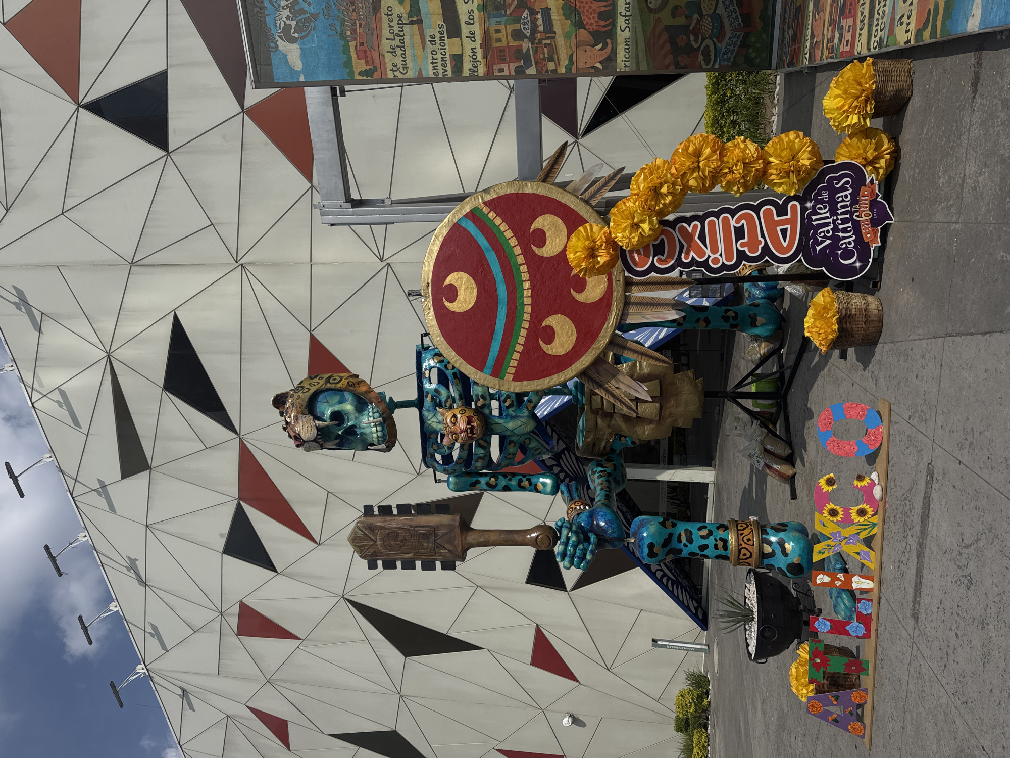 The image depicts a decorative installation in Atlixco, a city in the state of Puebla, Mexico. The main subject is a large, vividly colored skeleton figure, likely representing an Aztec warrior or deity, as suggested by the shield and club it wields. The skeleton figure is adorned with intricate patterns and designs. A sign with "Atlixco" and "Valle de Catrinas" (Valley of Catrinas) is placed nearby, indicating a Day of the Dead or similar festival. Marigolds, a traditional flower for Day of the Dead, are used as decorations. A vertical sign spelling "MEXICO" in colorful letters is also present. The background includes a building with a geometric pattern. The image is taken in an outdoor setting, likely a street or plaza.