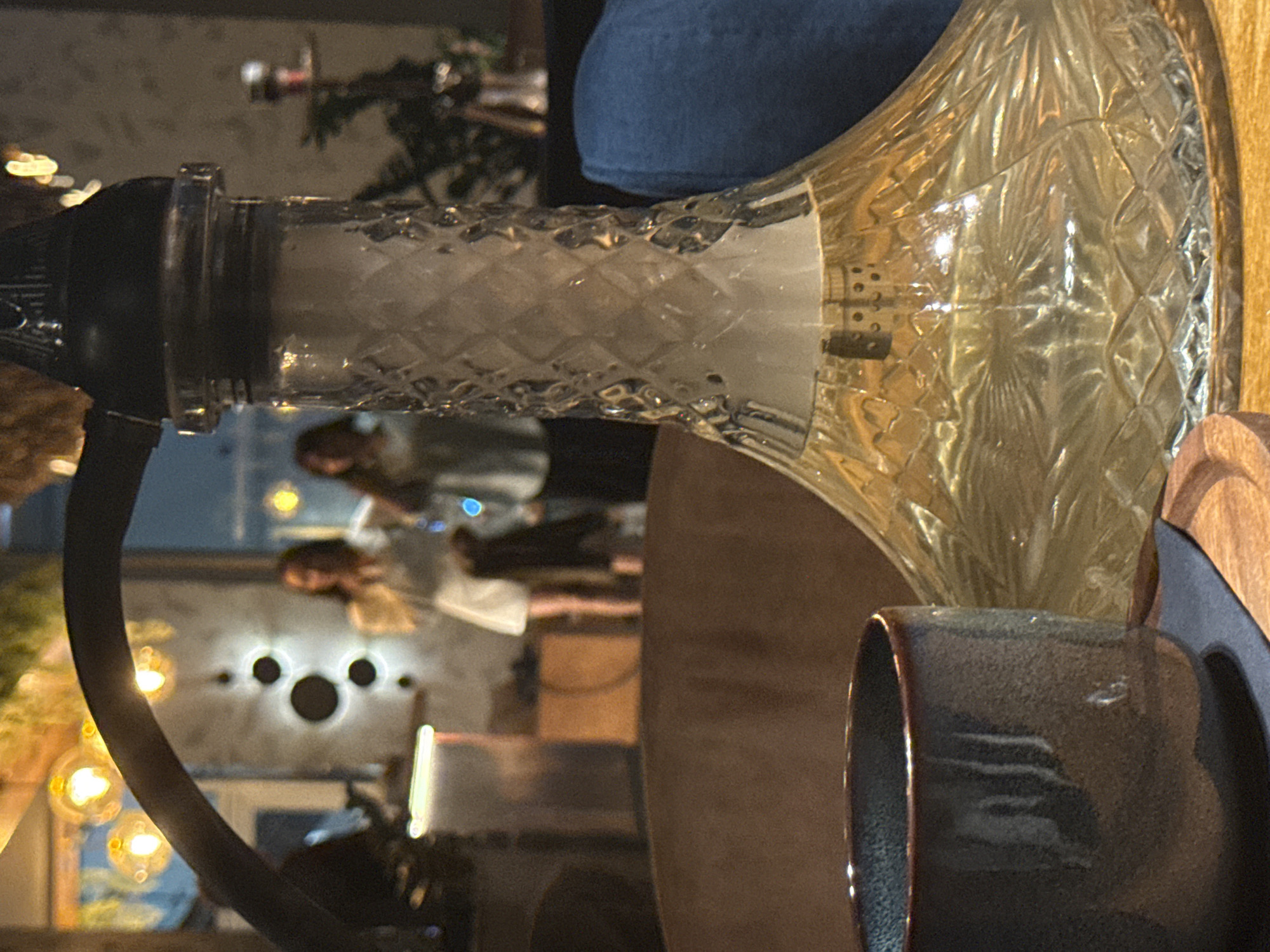 The image depicts a close-up of a hookah with a glass base filled with a yellow liquid. The base has a diamond-like textured pattern. In the foreground, to the left, is a dark, textured cup. In the background, out of focus, two women are visible in a dimly lit indoor setting. One woman wears a white top and skirt, and the other wears a light-colored top and dark pants. Decorative lights are visible in the upper background. The overall scene suggests a lounge or bar environment. The location is identified as Bratislava, Slovakia.