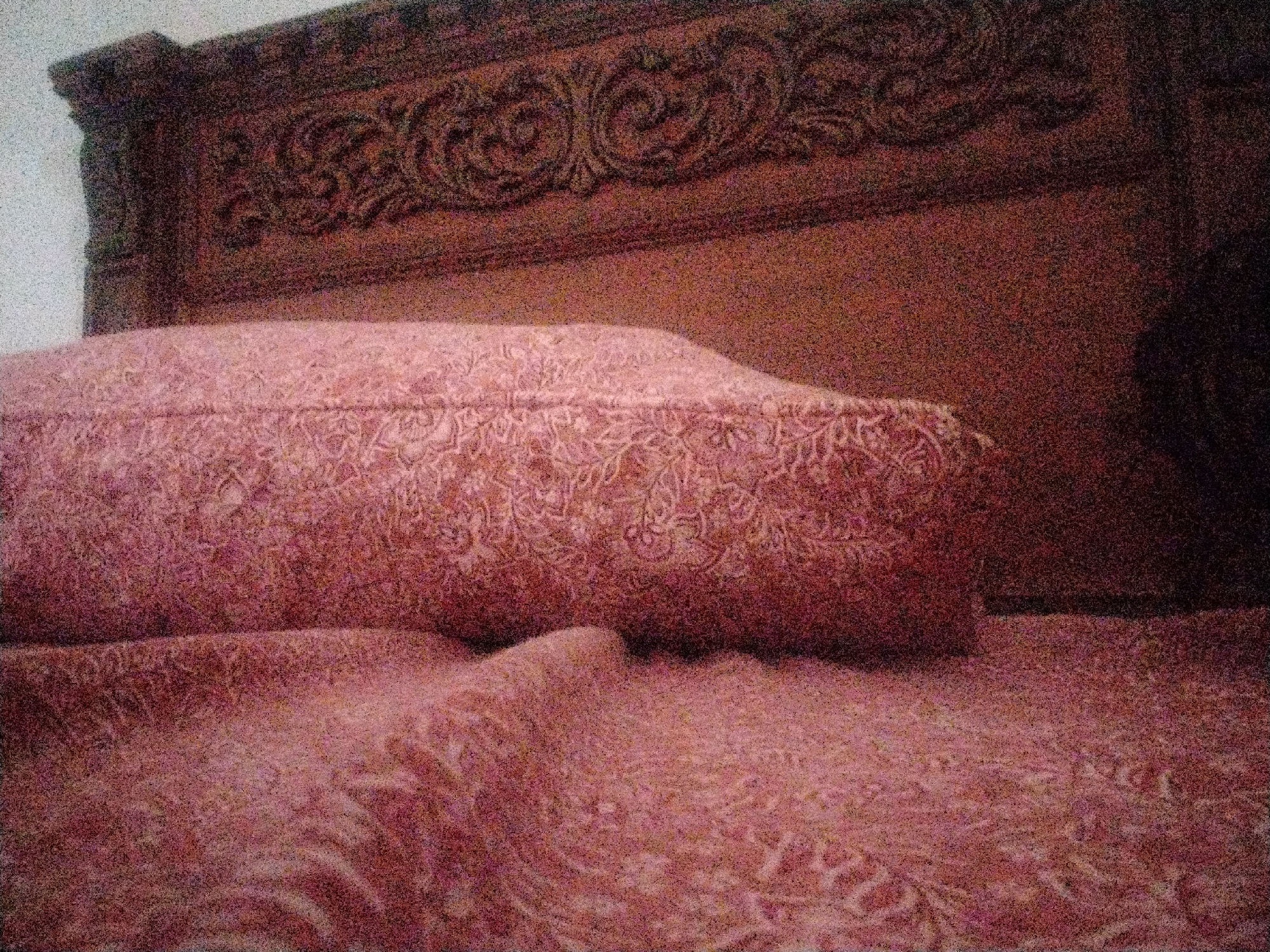 The image shows a close-up, slightly blurry view of a bed. The bed has a dark, ornate wooden headboard with intricate carvings. A pillow, covered in a pink fabric with a patterned design, rests against the headboard. The bed is also covered with a matching pink patterned sheet or blanket. The lighting in the image is dim, and the overall impression is of a bedroom interior. The location is Unknown, Unknown.