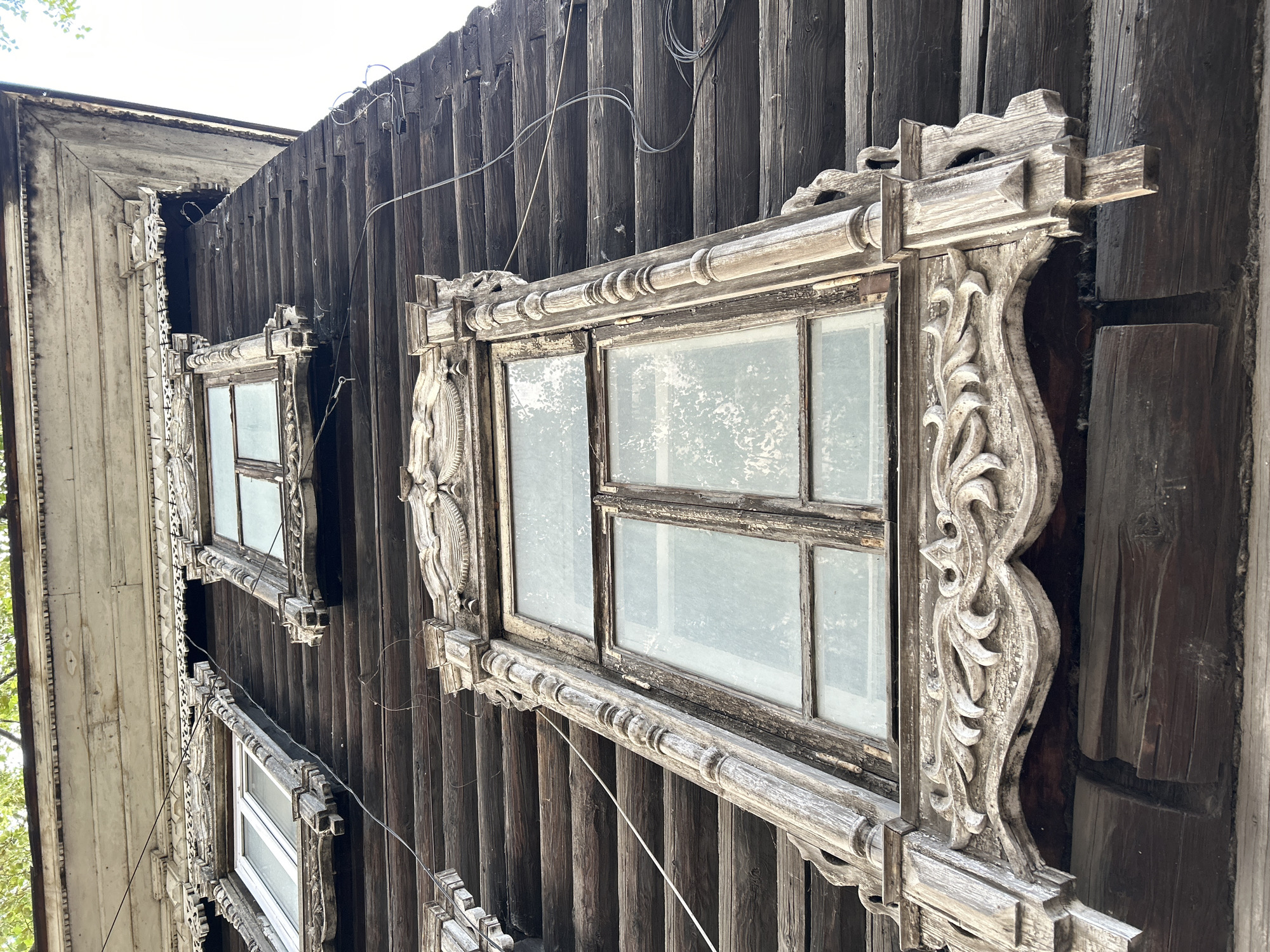 A low-angle, close-up shot captures the weathered, dark wooden exterior of an old log building in Tomsk, Russia, likely a historic residential structure. The focal point is a large, ornate window frame on the second story, featuring intricate, carved wooden detailing in shades of faded white and grey. The window itself has multiple panes, some of which are dusty and smudged. Above this main window, two smaller windows are visible, also with decorative, carved wooden surrounds and multiple panes. The building's construction is evident from the visible horizontal logs, showing signs of age and wear. Several thin wires, possibly for electricity or telecommunications, are strung across the facade, adding to the sense of age and an outdoor environment. The sky is bright and overcast, suggesting daytime. The overall impression is one of historical architecture in a state of gradual decay.