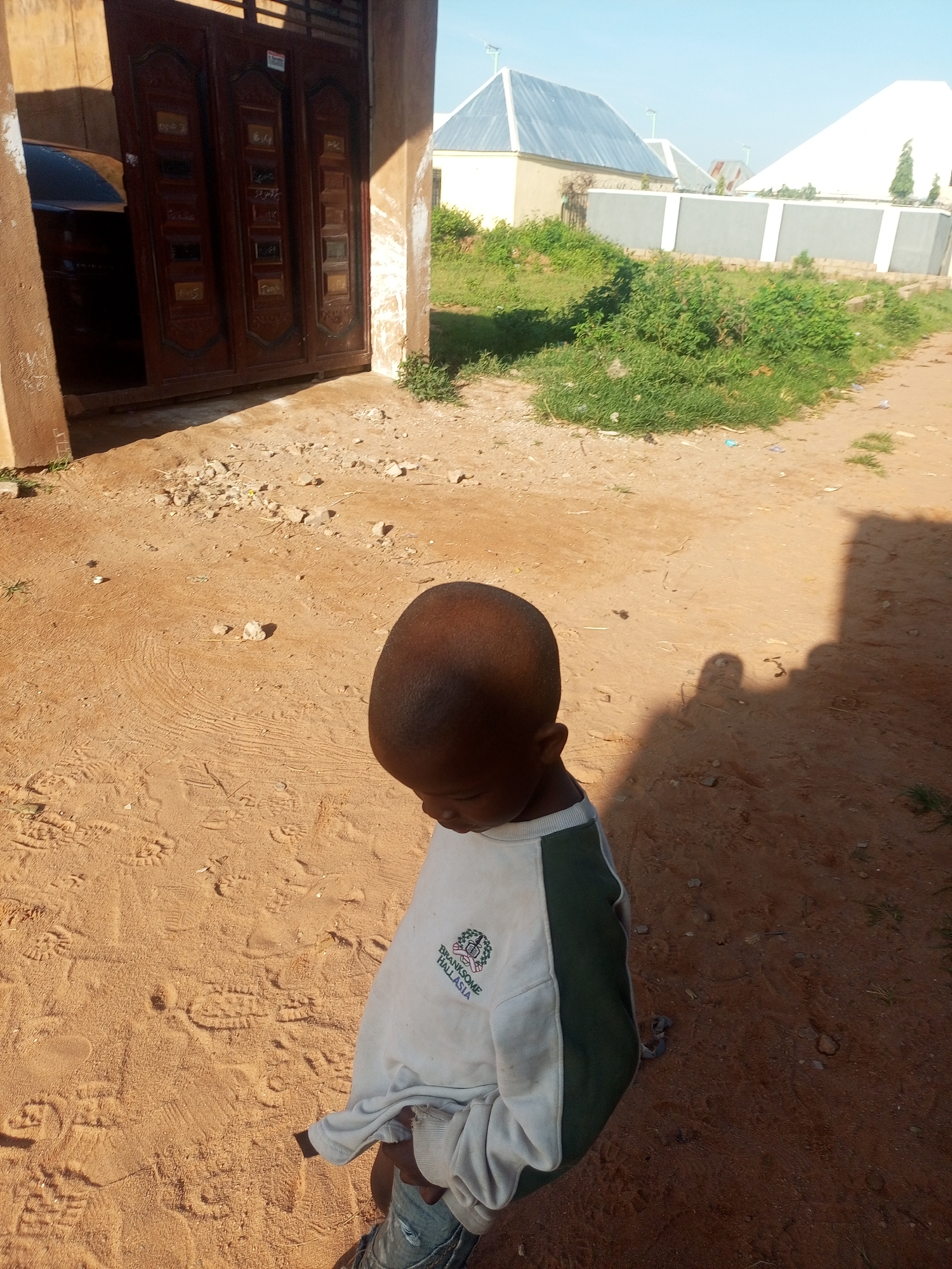 A young child, with dark skin and a shaved head, stands in a sandy outdoor area. The child is wearing a light-colored sweatshirt with dark green panels on the sides and a logo on the front, along with denim shorts. The ground is covered in fine, orange sand with visible footprints and small stones. To the left, a portion of a building with large brown double doors is visible, and a dark-colored car is partially parked behind the doors. In the background, there are several houses with distinct roof designs and a grey wall. The scene appears to be in a residential area with a clear blue sky overhead. The location is Tempure, Nigeria.